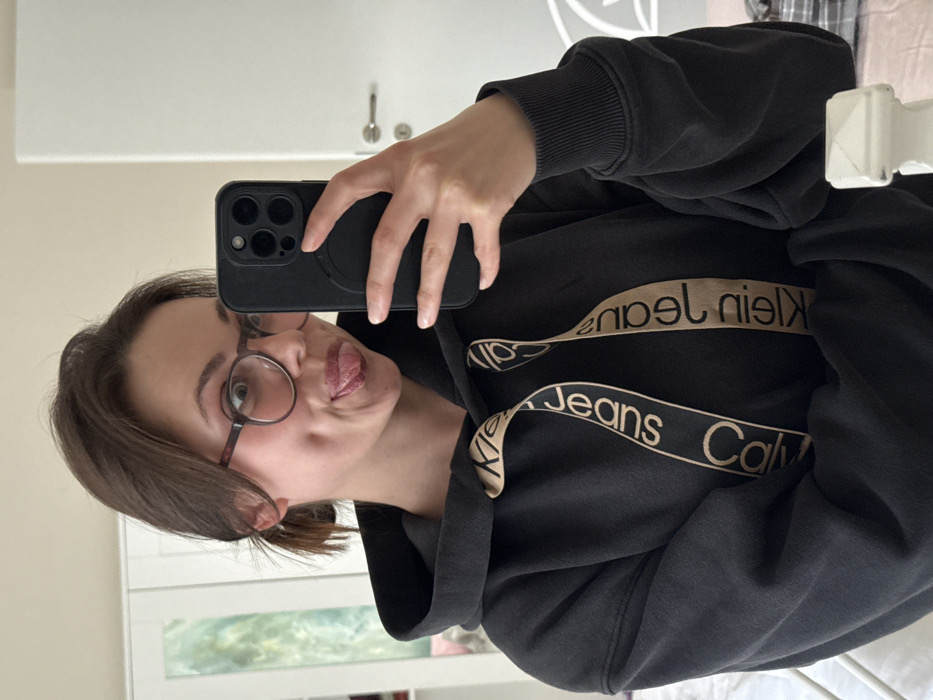 An indoor mirror selfie depicts a young adult female located within a private residence in Scafati, Italy. The subject, positioned centrally, is making a playful expression with her tongue extended, wearing round-framed eyeglasses with dark rims, and has dark brown hair pulled back. She is clad in a dark-colored (likely black or dark grey) hooded sweatshirt featuring beige drawstrings with the repeating "Calvin Klein Jeans" logo in dark lettering. Her right hand holds a dark-colored smartphone with a prominent triple camera array and a dark case, capturing her reflection. The background reveals elements of a room, including a light-colored wardrobe with reflective panels on the left, a white door with a silver handle on the right, and parts of a white, possibly metal, bed frame visible in the lower corners. The walls are a light, neutral color.