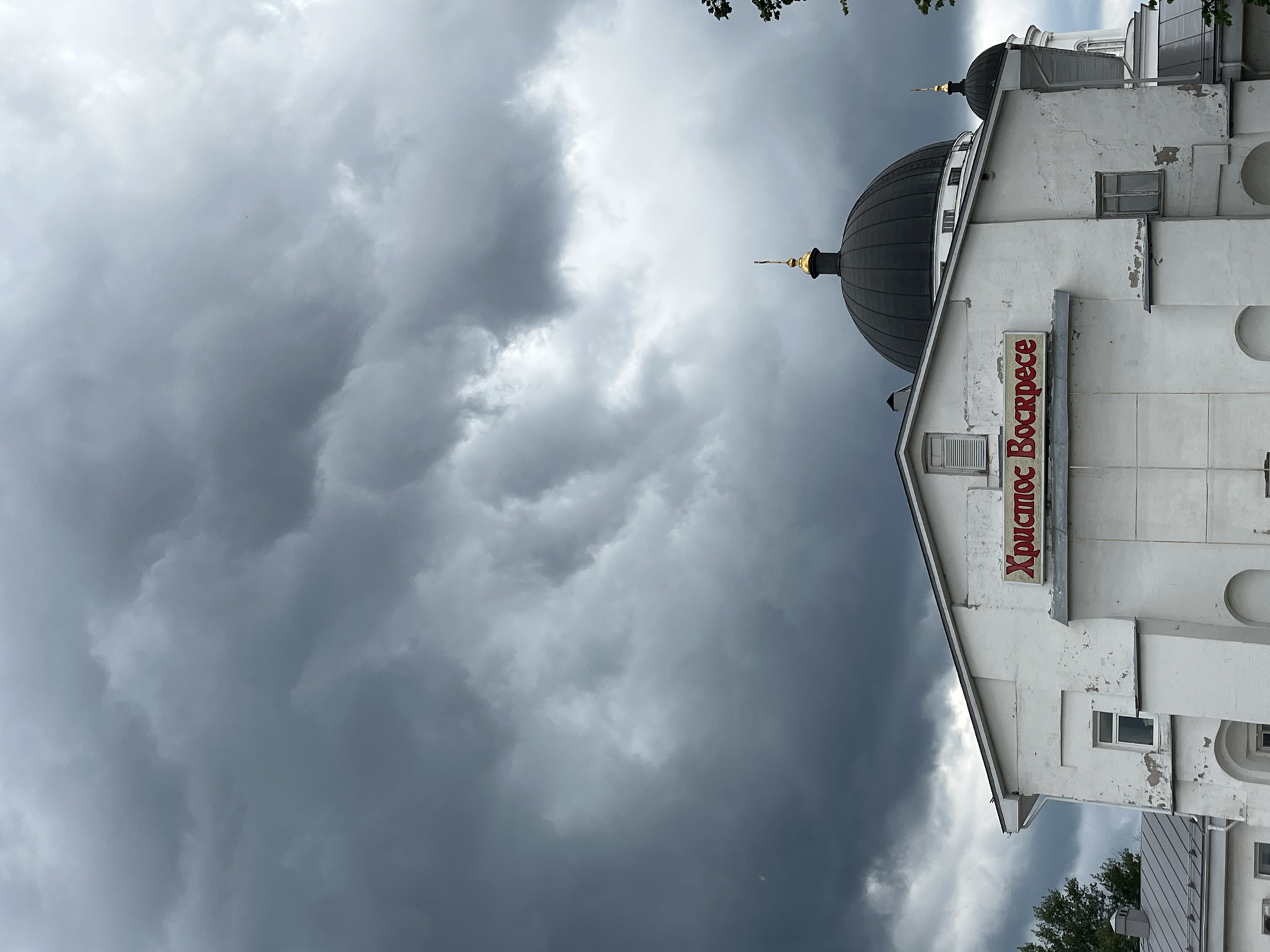 The image shows a close-up view of the white facade of a building, likely a church, with a dark dome against a dramatic, cloudy sky. A red sign with Cyrillic text that reads "Христос Воскресе" (Christ is Risen) is prominently displayed on the building's upper section. The building exhibits signs of age, with peeling paint and some architectural details suggestive of Russian Orthodox style. The dome, topped with a golden cross, is a focal point, as are the dark, turbulent clouds overhead, which create a somber atmosphere. The lower part of the building has windows and arched openings. The scene is set in Nizhny Novgorod, Russia.