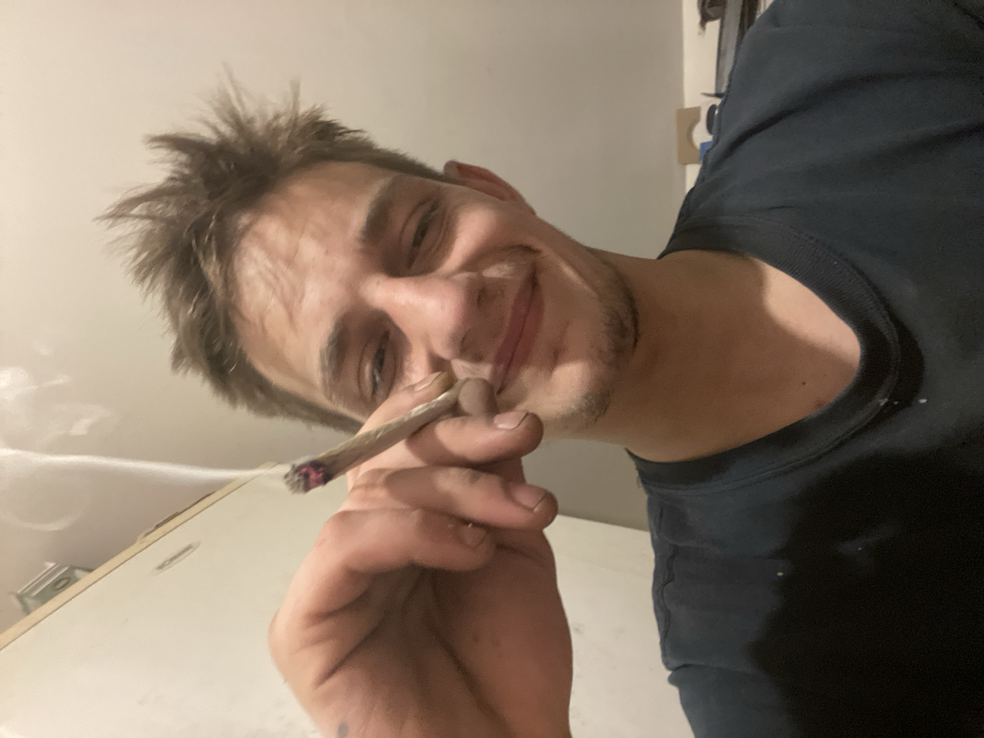 A young adult male, with light brown, spiky hair, and a faint mustache and stubble, is pictured from the chest up, looking towards the camera with a slight smile. He is holding a lit cigarette between the thumb and index finger of his left hand, with a trail of smoke rising. He is wearing a dark blue t-shirt. The background is a plain, light-colored wall with some indistinct objects visible in the upper right corner. The lighting suggests an indoor setting, possibly a room.