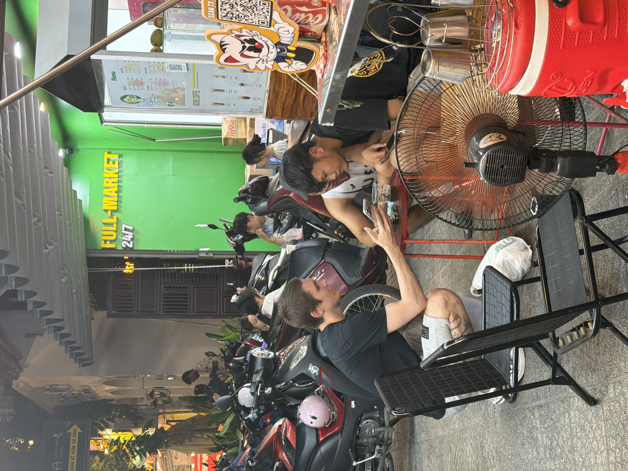 The image shows an outdoor cafe scene in Đà Nẵng, Vietnam. Several people are seated at tables, with motorcycles parked in front of the cafe. A man in a black shirt sits with his arm raised in conversation with another man next to him. A third man is using a mobile phone, possibly using the cafe's WiFi. The cafe has a green wall with the words "FULL-MARKET 24/7" in yellow. The menu is displayed with a cartoon cat graphic. A large red cooler is positioned near a fan. The image also shows a street scene with a sign pointing to a hotel, "Mouretou HOTEL ĐÁNG KỂ VIÊM, ĐÀ NẴNG."