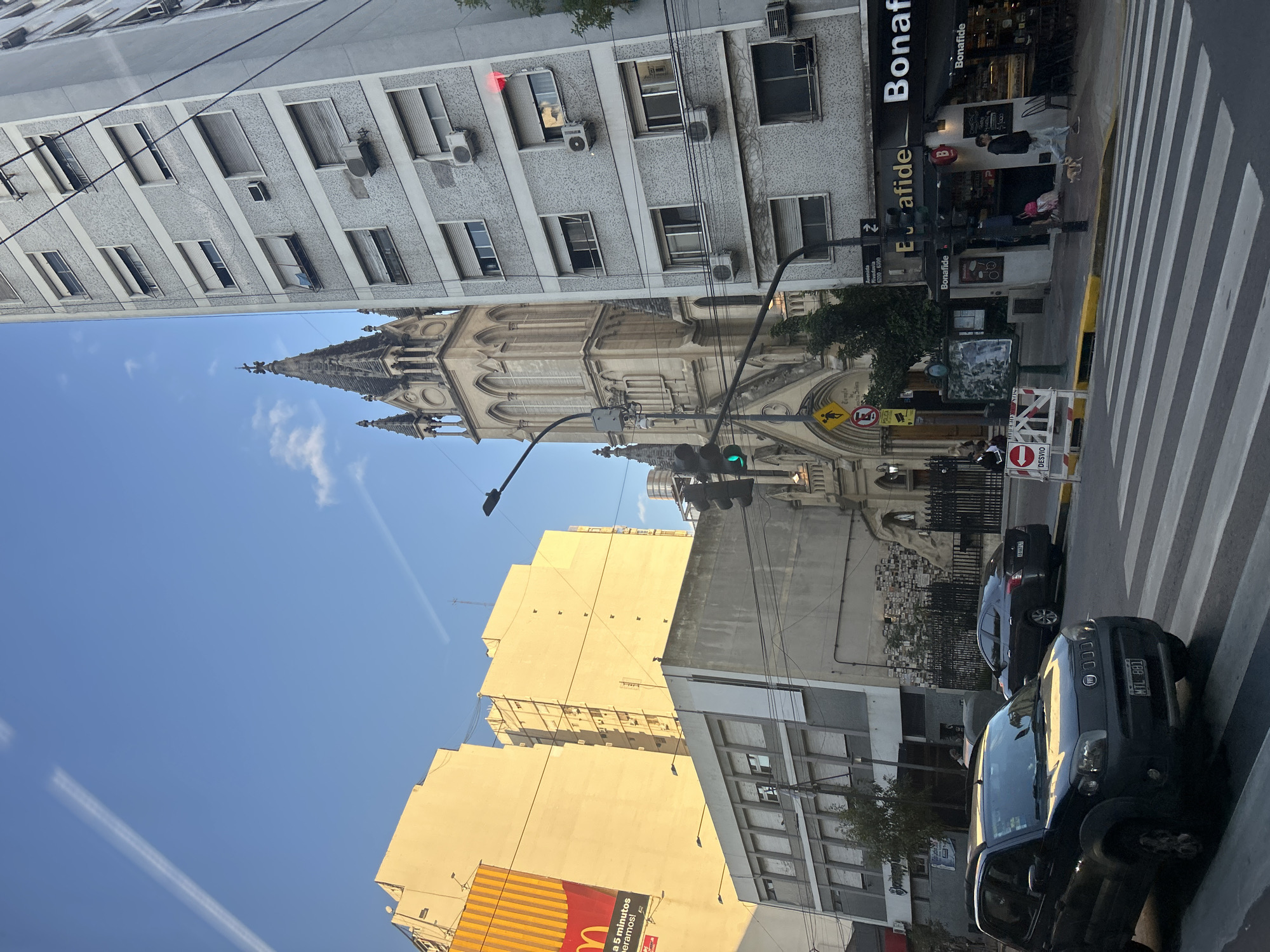 The image depicts a street scene in Buenos Aires, Argentina, featuring the neo-Gothic Basílica del Santísimo Sacramento (Basilica of the Most Holy Sacrament) towering between buildings on the left side of the frame. Several buildings are visible, including one with a Bonafide cafe at street level. A McDonald's sign appears on a building further down the street. Two cars are parked at a crosswalk, and pedestrians are near the cafe. A traffic light is in the center of the frame with a green light visible. Street signs and barriers are also present. The sky is blue with wisps of clouds.