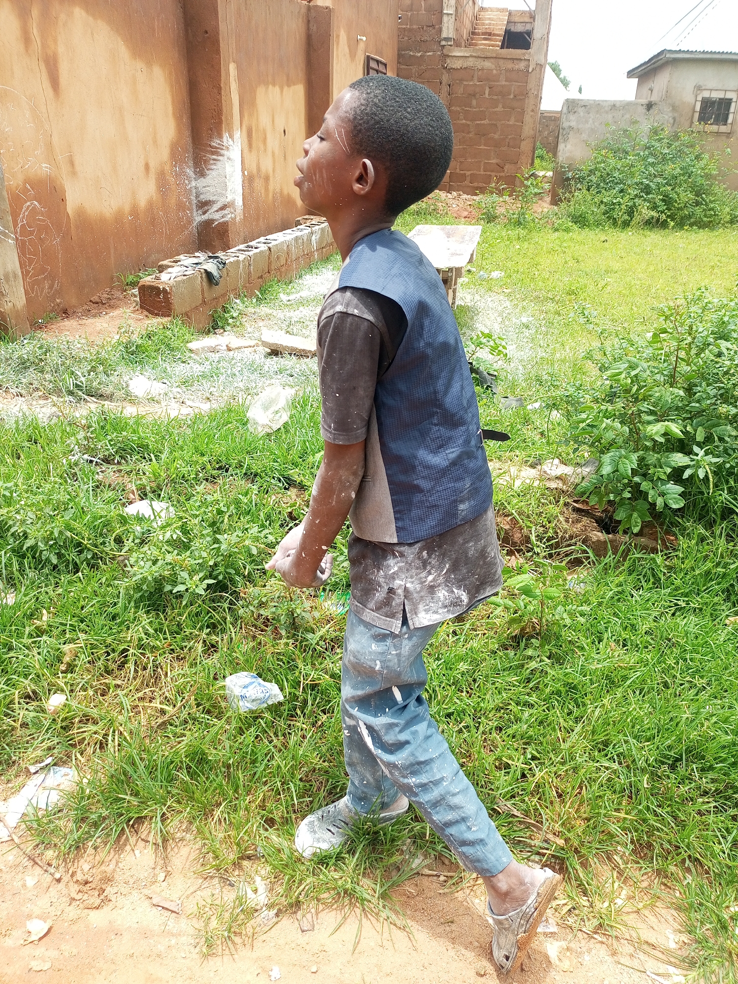 The image depicts a young boy standing outdoors, facing left, in a dusty and overgrown area. He is wearing a dark grey t-shirt, a blue patterned vest, and faded blue pants. His clothing and skin are smudged with white material, likely paint or plaster. His face is turned upwards, with his eyes closed or looking upwards. The background shows a weathered, reddish-brown wall with some white splatters and chalk-like drawings. A low wall made of concrete blocks is visible in front of the wall. The ground is a mix of dirt and sparse, overgrown green grass, with some scattered debris including plastic bags and white residue. In the distance, partially constructed buildings with exposed brickwork and a metal roof are visible, along with dense green foliage. The overall setting appears to be a construction site or an area undergoing renovation in a semi-urban or rural environment. The provided information suggests this location is in Tempure, Nigeria.