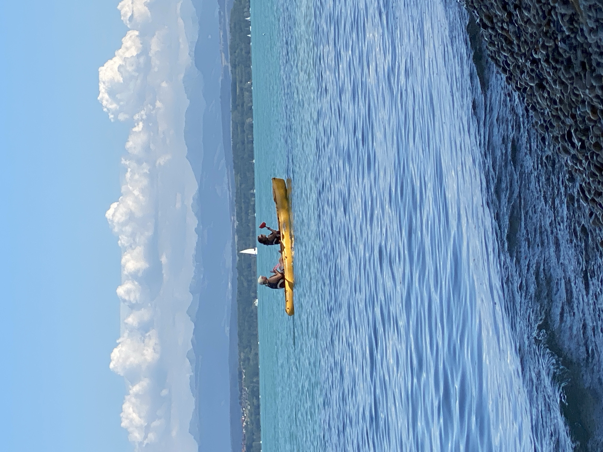 In the city of Cortaillod, Switzerland, two individuals are seen kayaking on a tranquil, turquoise lake under a bright blue sky filled with fluffy white clouds. A distant mountain range, shrouded in a hazy blue, forms the horizon. The foreground of the image is occupied by the rippling surface of the lake, transitioning into a pebbled shoreline. The two kayakers, positioned in a yellow double kayak, are actively paddling. One individual, in the front, is wearing a hat and a life vest, while the person behind them, also wearing a life vest, holds a red paddle. Several sailboats are dotted across the lake, and a sailboat with a white sail is visible in the middle distance. A green, treed landscape with scattered buildings lines the far shore of the lake.