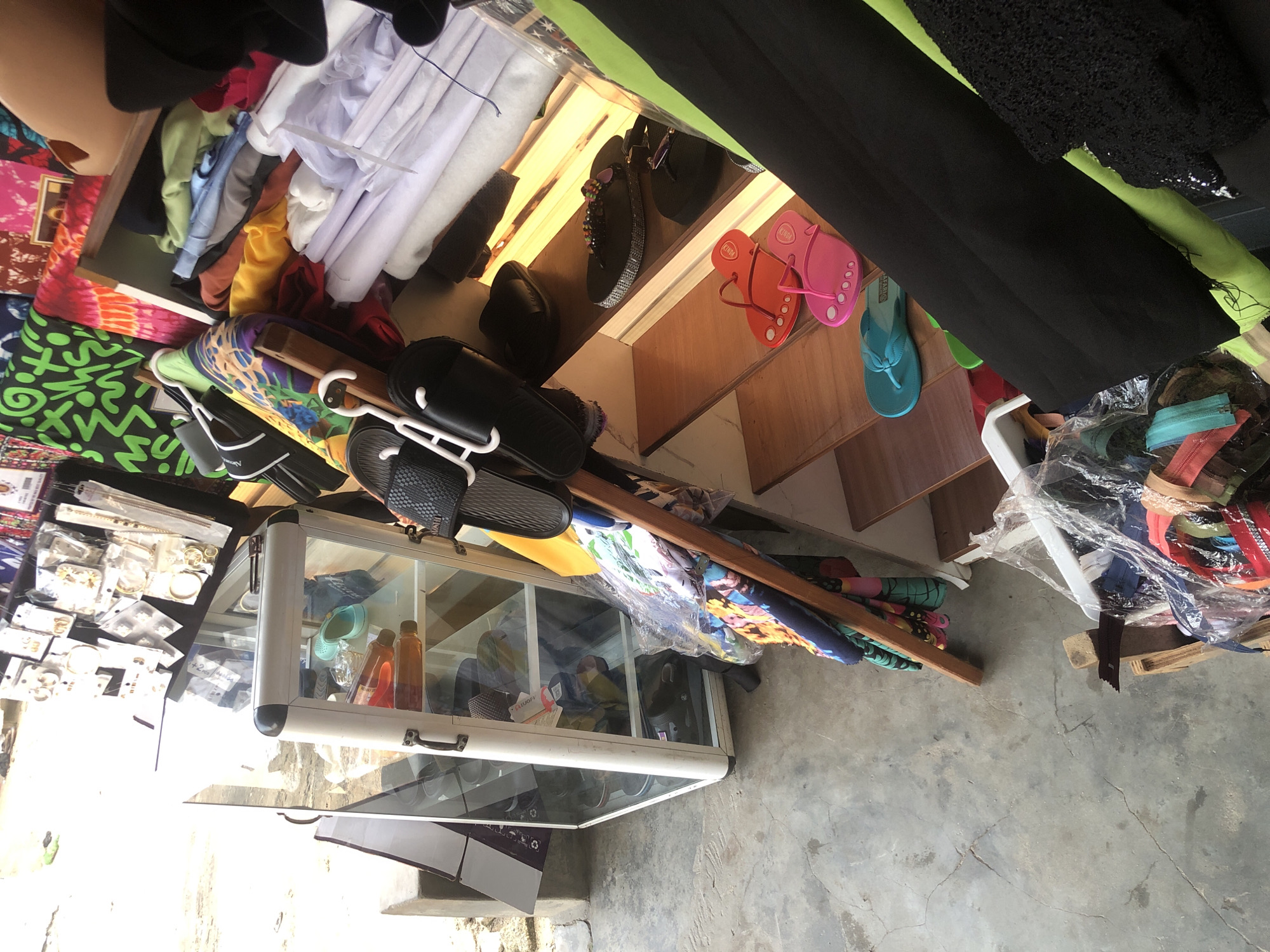 This image depicts a detailed view of an outdoor market stall or a small shop in the city of Ado, Nigeria, under bright daylight conditions. No individuals are visible within the frame.
The scene is characterized by a grey, cracked concrete floor in the foreground and left side. A white-framed glass display cabinet stands on the left, showcasing a single teal sandal and two bottles of amber-colored liquid on its top shelf, with other dark footwear vaguely visible on lower shelves. A concrete block and a partially obscured purple and white cardboard box are positioned to the cabinet's left, beyond which a brightly lit outdoor area is visible.
In the central area, a rustic wooden stand displays multiple pairs of black slides/sandals hanging on white plastic hangers. Colorful patterned fabrics are draped or folded beneath these items.
The right side of the image features a multi-tiered wooden shelving unit. The upper shelves are laden with stacks of folded garments in various colors including light green, blue, brown, yellow, and white. Above these, brightly patterned fabrics (green, tie-dye, red) are arranged, and a dark board displays numerous pieces of packaged gold-colored jewelry, including earrings and necklaces. The lower shelves exhibit several pairs of sandals and flip-flops: two dark flat sandals (one embellished with colorful beads), a pair of orange flip-flops, a pair of pink flip-flops, and a pair of light blue flip-flops.
The bottom right foreground contains folded black fabric, a black lace-patterned garment, and a clear plastic bag filled with an assortment of colorful zippers, resting on what appears to be a wooden crate or pallet.