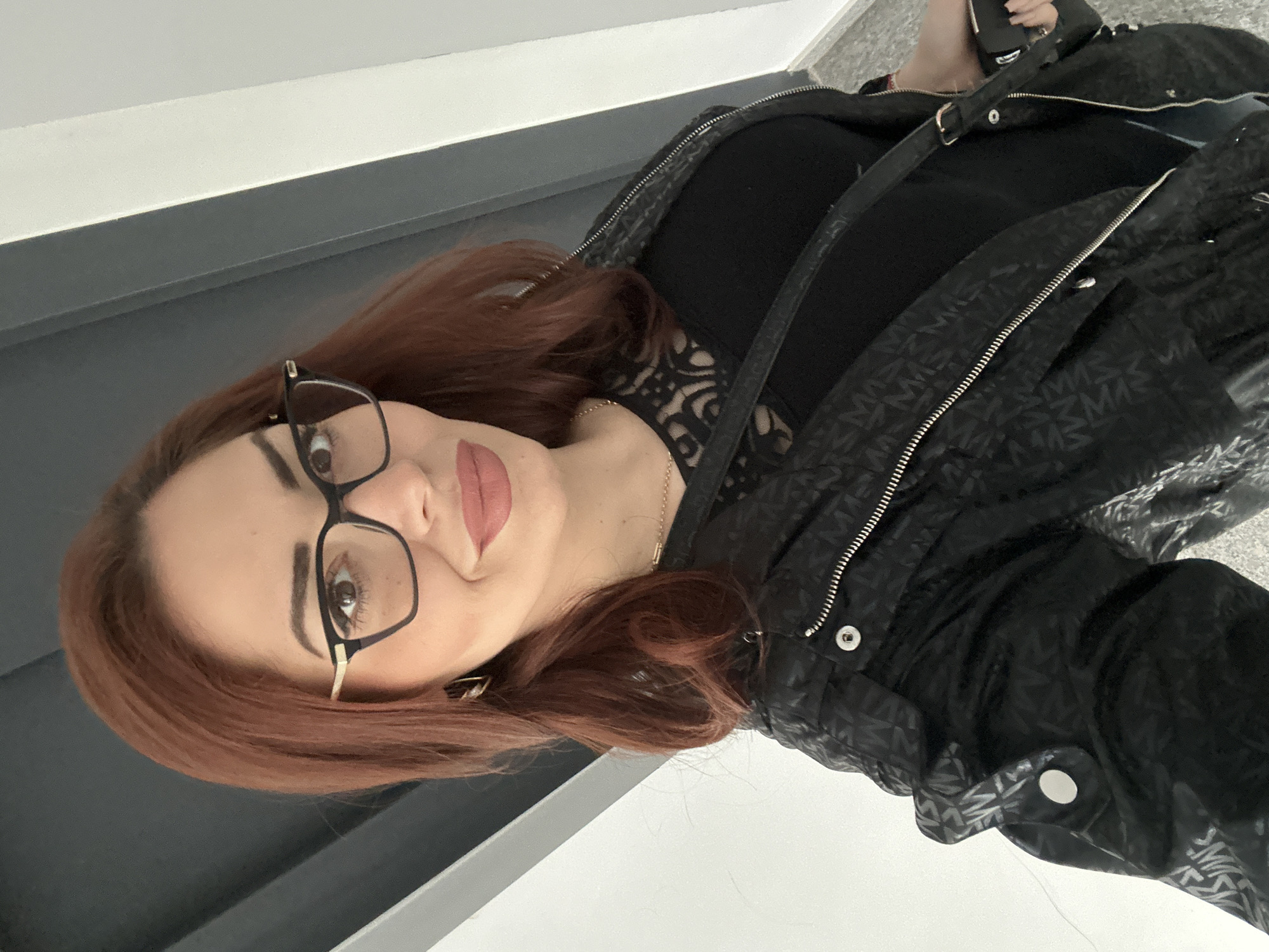 A woman with auburn hair is wearing black-framed glasses and a black jacket with a subtle pattern. She has fair skin and is wearing lipstick. She is standing in what appears to be an indoor location. The environment includes white and grey surfaces, possibly walls or floors. The specific location in Sofia, Bulgaria cannot be determined from the image.