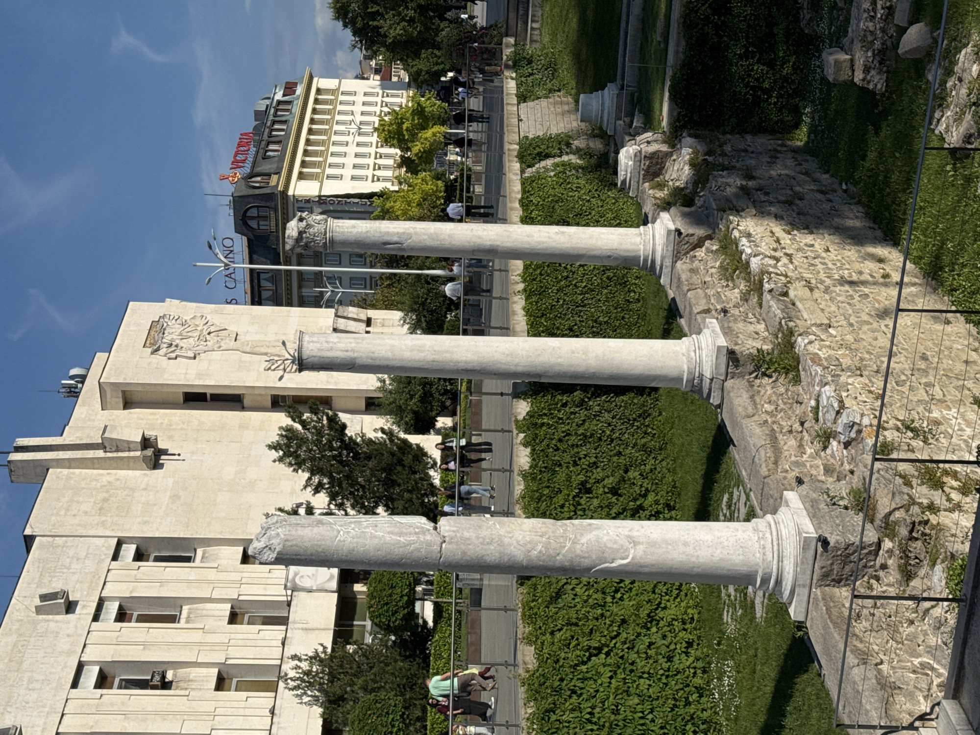 The image shows a view of the ancient Roman ruins in Plovdiv, Bulgaria. Three partially reconstructed columns stand along a pathway. Behind the columns, a stone wall runs along the pathway. A metal fence is partially visible, separating the ruins from a grassy area. People can be seen walking along the pathway. In the background, modern buildings are visible, including a building with a prominent "VICTORIA" sign. The sky is blue, and the overall lighting suggests a sunny day. The buildings appear to be of typical urban construction.