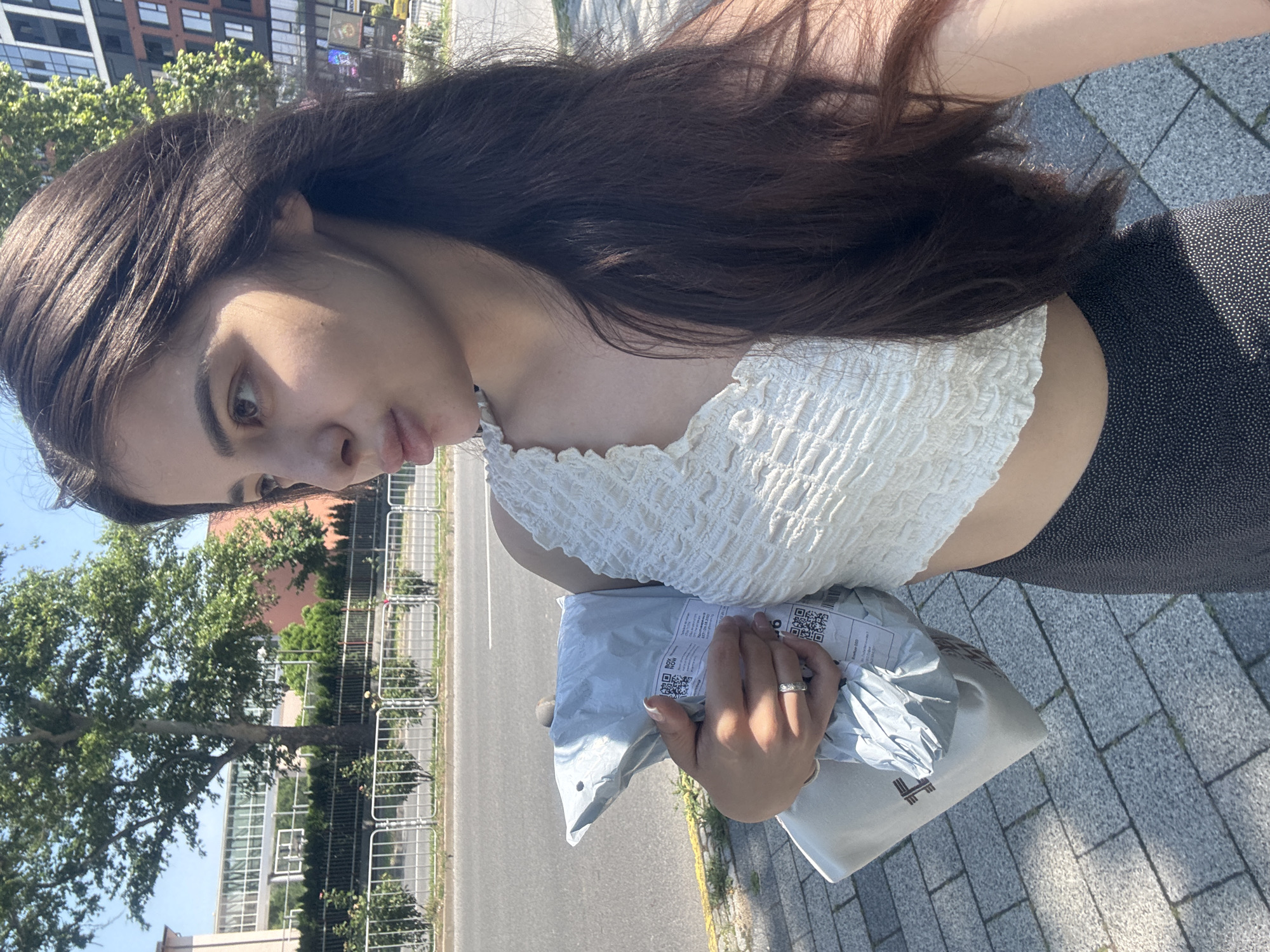 A young woman is captured in a mid-shot portrait on a sunny day in Plovdiv, Bulgaria. She stands on a grey paver sidewalk, holding two packages in her left arm and hand. The primary package is a light grey poly mailer featuring white labels with QR codes and text in Cyrillic script. Beneath it, a second, lighter-colored bag with a dark brown logo is partially visible. The woman wears a white, textured, one-shoulder crop top and dark bottoms with a small white polka dot pattern. A silver-toned ring is visible on her left ring finger, and her fingernails are manicured with white tips. Her long, dark brown hair falls over her shoulders. Her face is turned to her right, with her lips slightly pursed, and the right side of her face is brightly illuminated by direct sunlight, casting shadows. In the background, an asphalt road is visible to the left, separated from the sidewalk by a white metal pedestrian barrier. Behind the barrier are green shrubs, small rose bushes, and large deciduous trees. Further back, various buildings are visible: a modern light-colored structure with a skybridge to the far left, an orange-hued building, and a multi-story building with dark window frames to the far right. The sky is clear blue.