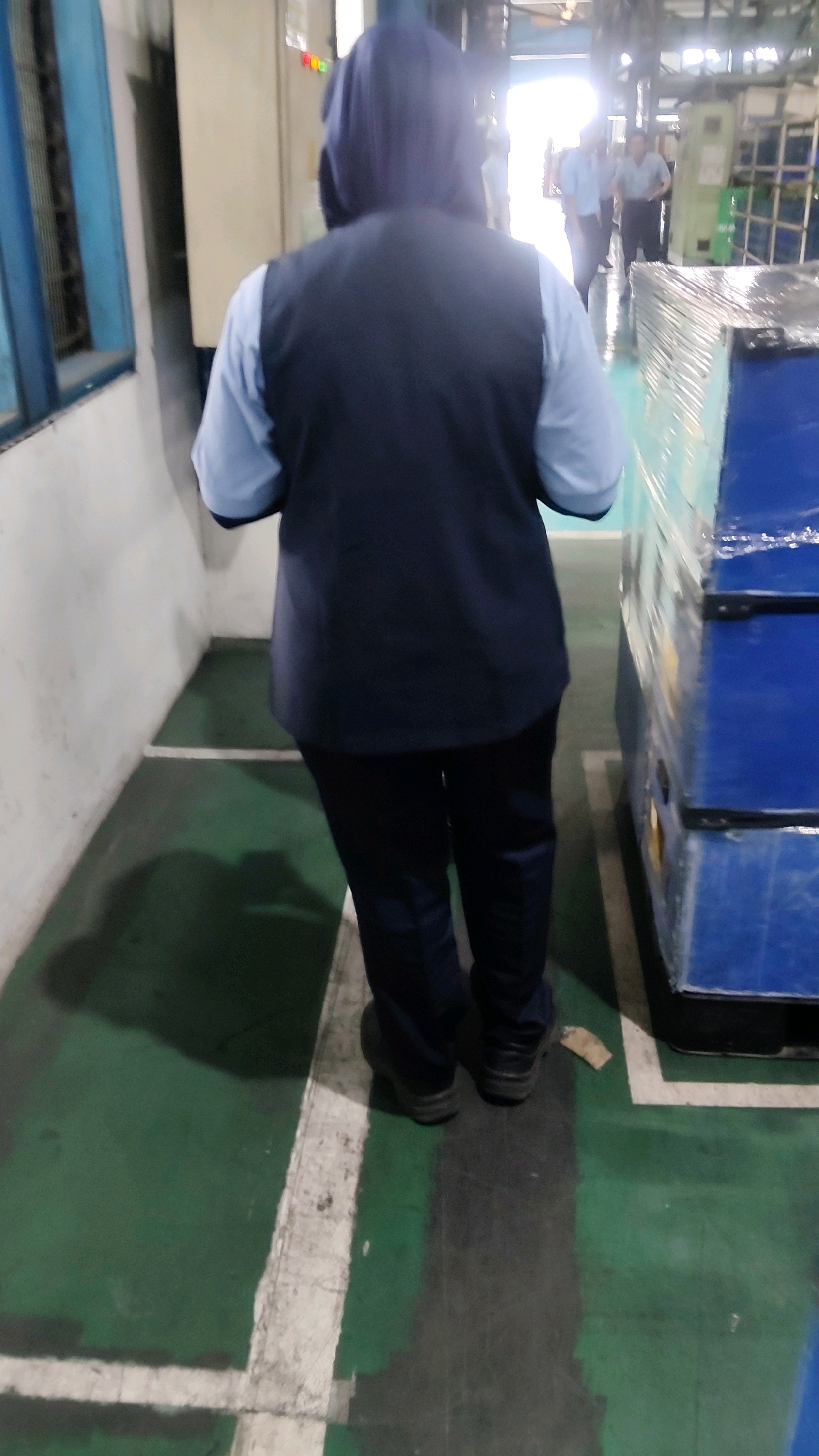 The image shows the back of a person wearing a dark blue vest over a light blue shirt and dark pants, with a dark blue hood covering their head. They are standing on a green floor with white lines, casting a shadow to their left. In the background, other individuals in similar uniforms are visible walking towards a bright light. To the right, a large blue object covered in clear plastic wrap is present. The environment appears to be an industrial or warehouse setting. The specific location in Mekarmukti, Indonesia, cannot be determined from the visual information provided.