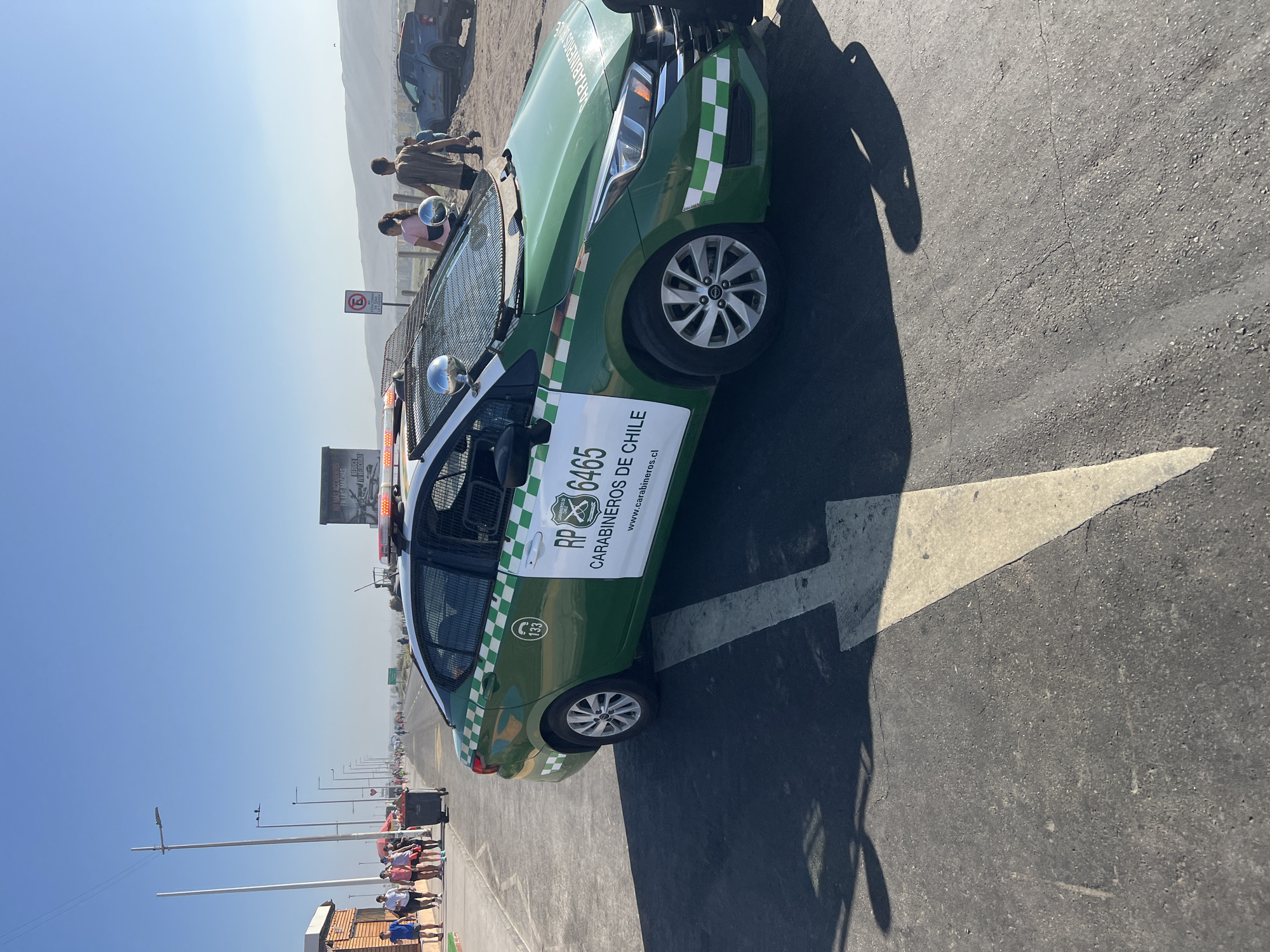 The image shows a Carabinero de Chile police car parked on a paved surface with a large white arrow painted on it. The car is green and white with checkered accents and displays the license plate "RP 6465" and the text "CARABINEROS DE CHILE." The car is parked on the side of a road. In the background, there are people walking along the side of the road, and buildings can be seen in the distance. There is a street sign. The sky is blue and clear. The location is in Arica, Chile. There is a sign on a truck in the background and on the police car about accidents and to reduce speed.