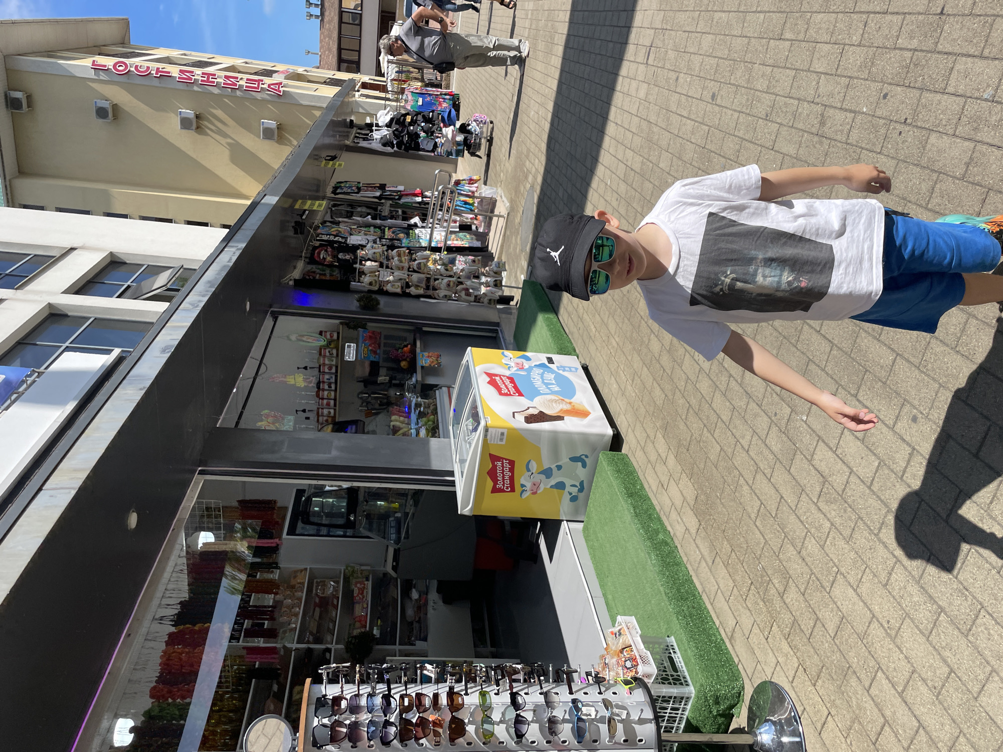 A young boy wearing a white t-shirt with a graphic print, blue shorts, and a black baseball cap with a white Jumpman logo is standing on a paved sidewalk in front of a store. He is also wearing sunglasses with green lenses. Behind him, there is a store with a display of sunglasses on a rotating rack to the left, and shelves stocked with various items inside the store. A white freezer with a cartoon cow and ice cream advertisements is situated outside the store entrance. Further down the sidewalk, a man in a grey t-shirt and pants is standing, and a building with Cyrillic signage that reads "ГОСТИНИЦА" (Gostinitsa, meaning "Hotel") is visible in the background. The scene appears to be a commercial street in Sochi, Russia, under a clear blue sky.