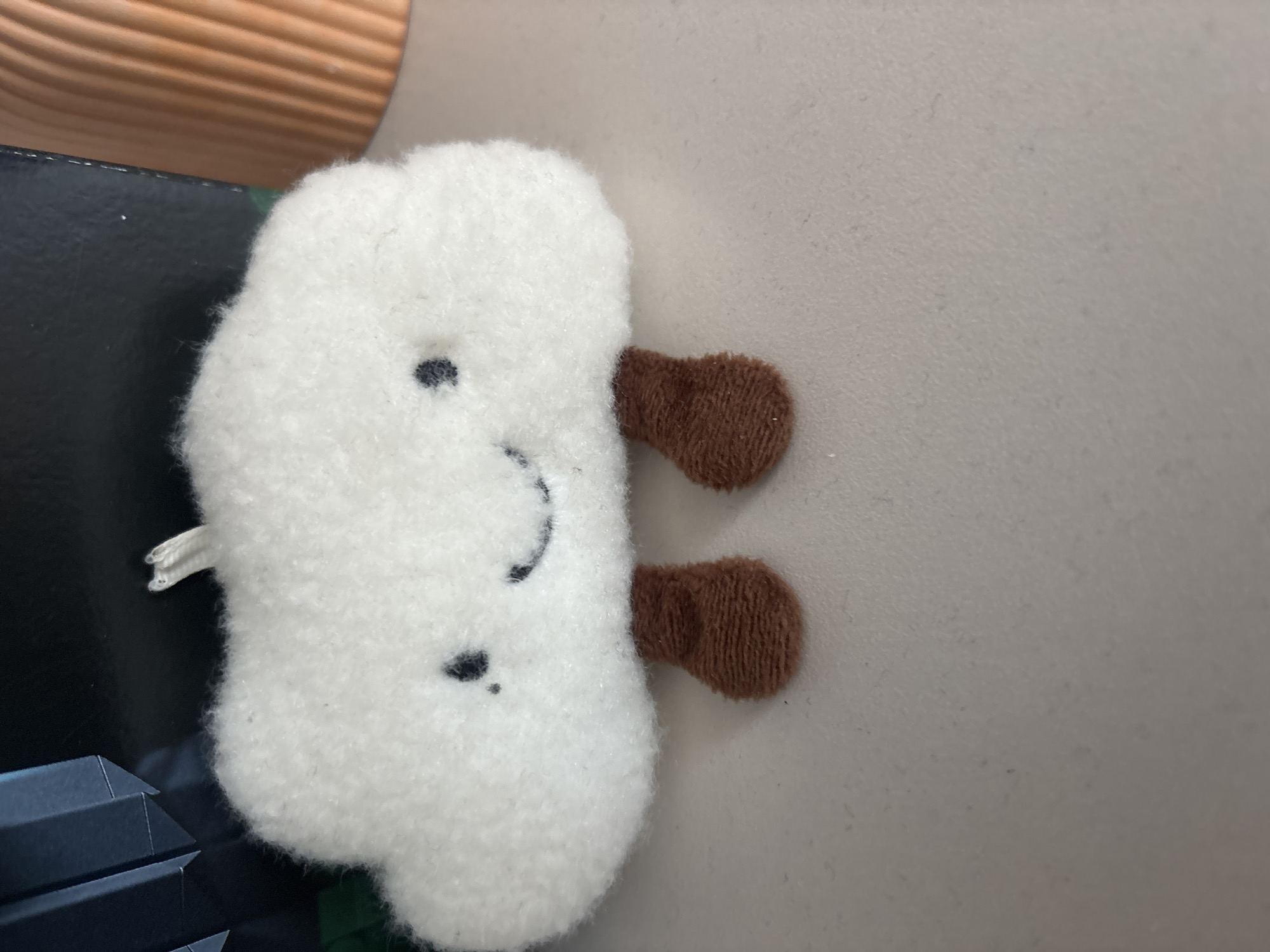 A close-up shows a small, white, plush toy with brown appendages, lying on a light-colored surface, next to what appears to be a dark-colored object. The toy has a simplified, embroidered face with two dots for eyes and a curved line for a mouth. It is difficult to discern the location of the scene.