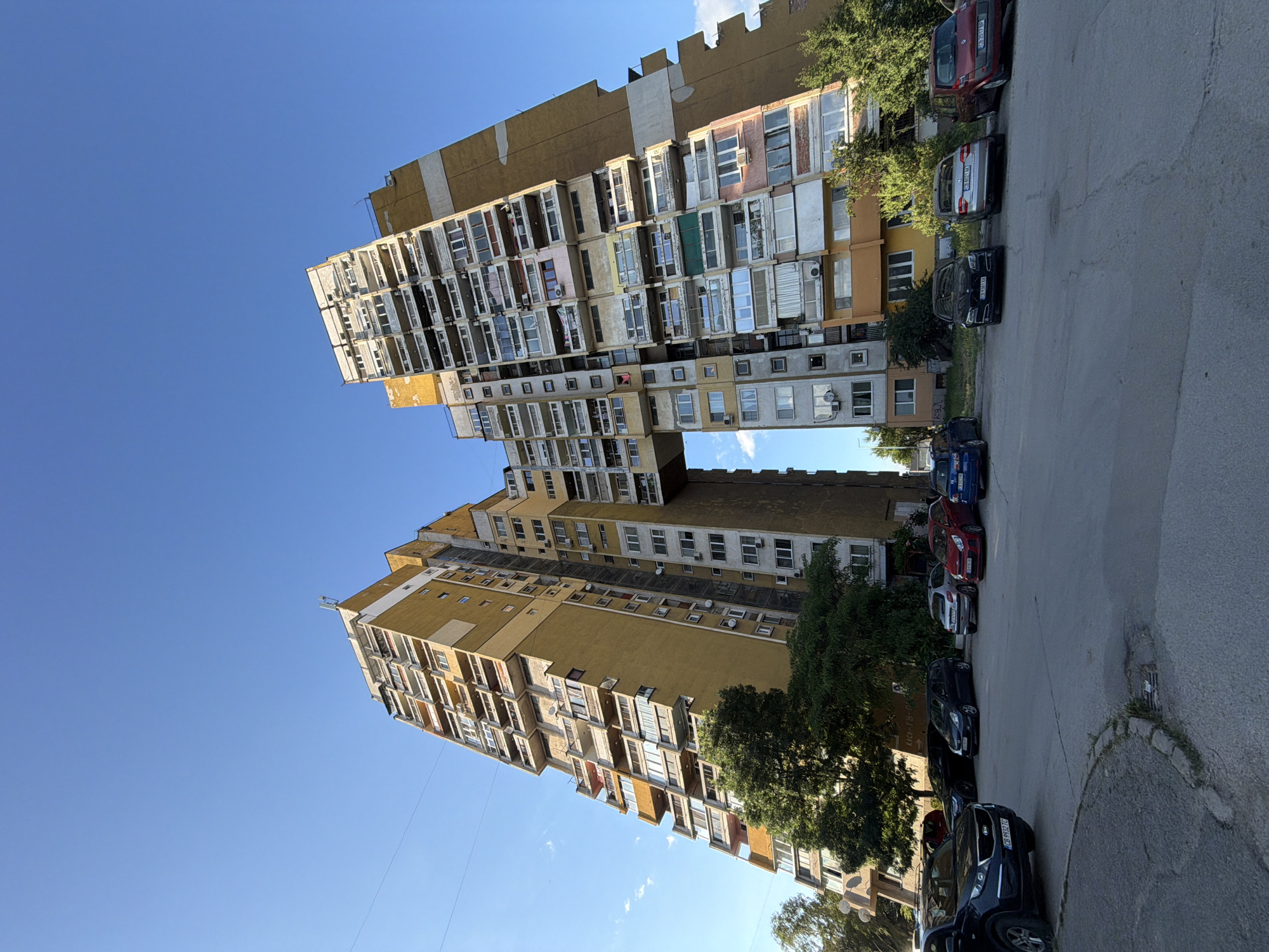 A wide-angle, low-angle shot captures a strikingly unusual Brutalist residential building complex in Sofia, Bulgaria, under a clear blue sky. The building, painted in shades of yellow and beige, is composed of two towering, multi-story structures that are angled towards each other, with a connecting bridge spanning the gap between them at a high level. The facades of both towers are densely packed with balconies, windows, and air conditioning units, suggesting a high population density within the apartments. Vegetation, including mature trees, is visible at the base of the building, with some branches reaching up towards the lower floors. In the foreground, a paved area appears to be a parking lot, with several cars parked in various positions. These include a black SUV in the left foreground, a black sedan, a dark gray hatchback, a silver sedan, a red hatchback, and a blue hatchback parked further down the row. The architecture of the building is characterized by its severe, geometric forms and the prominent use of concrete, typical of Brutalist style. The overall impression is one of imposing scale and unique architectural design.