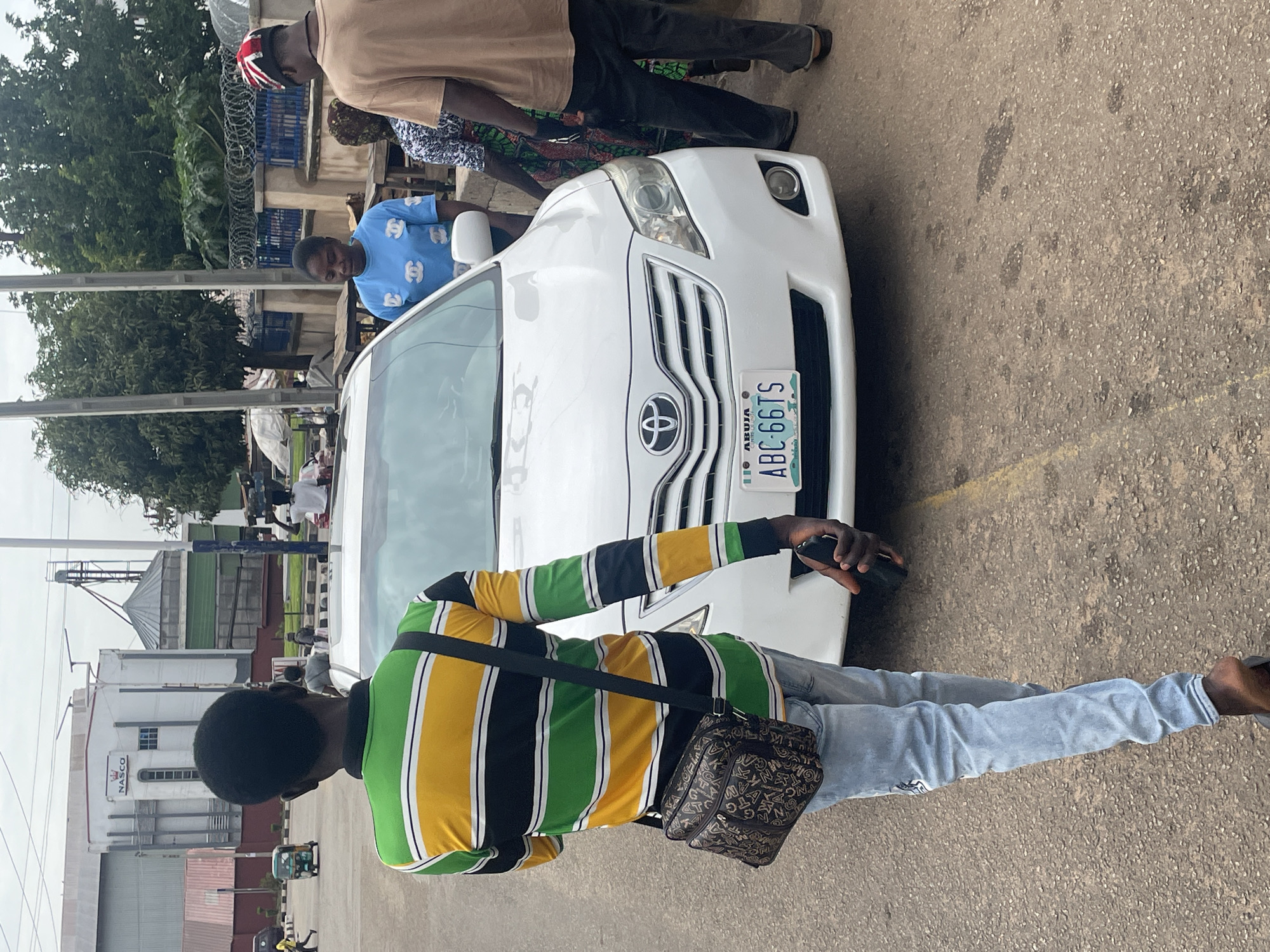 The image depicts a street scene in Jos, Nigeria, with a white Toyota Camry in the foreground. A young Black male, wearing a striped long-sleeved shirt and light blue jeans, walks away from the camera, carrying a dark brown shoulder bag and holding a black mobile phone in his right hand. Behind the car, a Black female in a blue t-shirt and another Black individual in a brown t-shirt are visible, alongside a third person partially obscured. The car's license plate reads "ABUJA ABC 66TS". In the background, a large industrial building with "NASCO" branding and grain silos are present, indicative of an industrial area. The ground is paved with asphalt, and a yellow line marks a lane divider. Trees and a fence with barbed wire are also part of the environment. The overall atmosphere appears to be a typical day in a city, with pedestrian and vehicular activity.