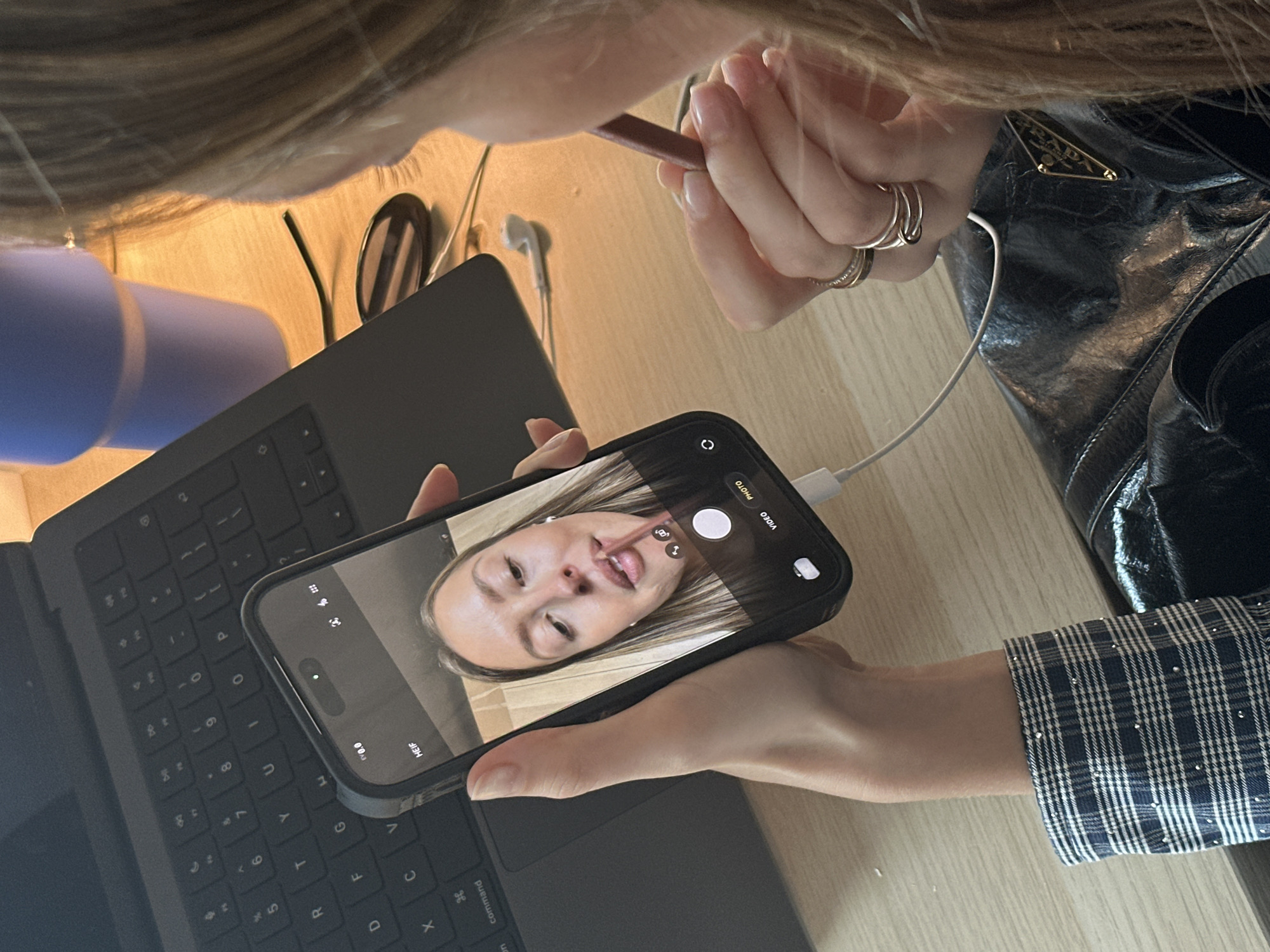The image shows a person using a brown lip liner and filming themselves on their phone. The phone is plugged into a MacBook keyboard. Next to the keyboard is a blue water bottle, wired earphones, and a black leather Prada bag. The person is wearing a blue and white checkered long-sleeved shirt. The location appears to be indoors.