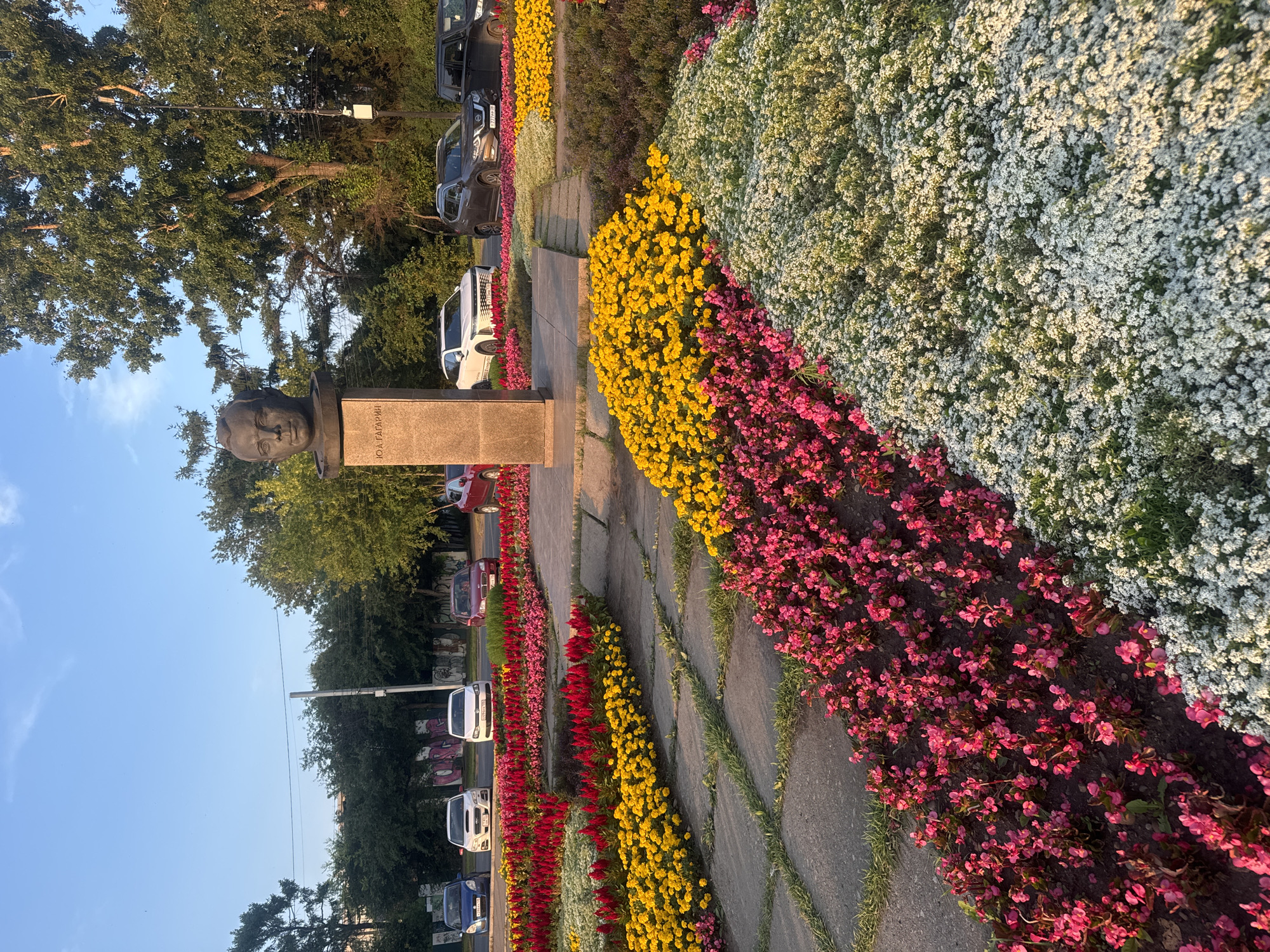 In Irkutsk, Russia, on a sunny day, a bust of Yuri Gagarin, with the inscription "Ю.А. ГАГАРИН" on its pedestal, stands prominently in a park. The monument is surrounded by a vibrant, multi-colored flowerbed featuring rows of pink, yellow, and white flowers. Paved pathways weave through the floral displays. In the background, several cars are parked along a street, with trees and a clear blue sky overhead. The image captures a peaceful, well-maintained urban landscape.