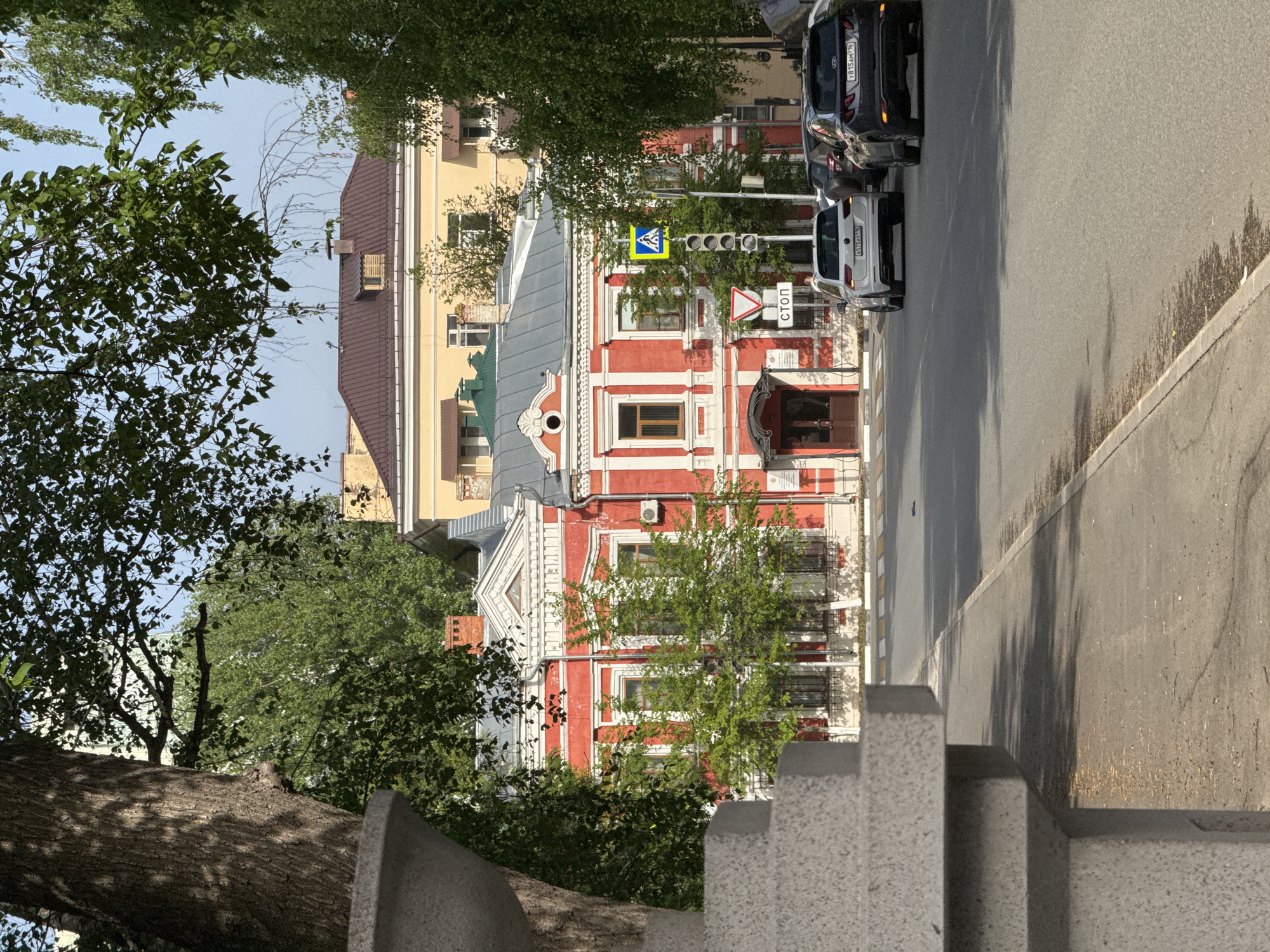 A street scene in Kazan, Russia, presents a red and white trimmed two-story building near a crosswalk with a pedestrian crossing sign and a stop sign in Cyrillic. Two cars are parked along the right side of the street. The buildings are partially obscured by trees, and the scene is viewed from an elevated position, overlooking a gray concrete barrier or ledge.