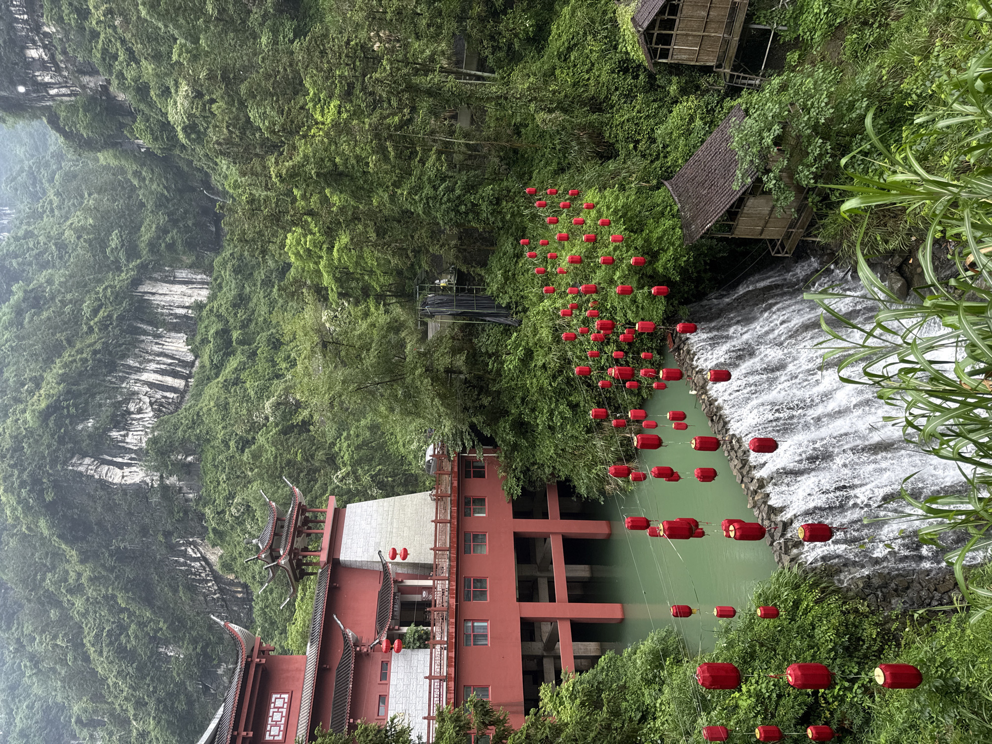The image shows a landscape in Yongding District, China, featuring a red building with traditional Chinese architectural elements. The building is constructed partially over a body of water, likely a river or pond, which has a greenish hue. Strings of red lanterns are strung across the water and through the surrounding vegetation. A waterfall cascades into the body of water, adding to the visual complexity of the scene. The area is heavily vegetated with lush green trees and foliage, contributing to a natural, forested environment. A small wooden structure or hut is visible further up near the waterfall. The background includes a cliff face covered in dense greenery.