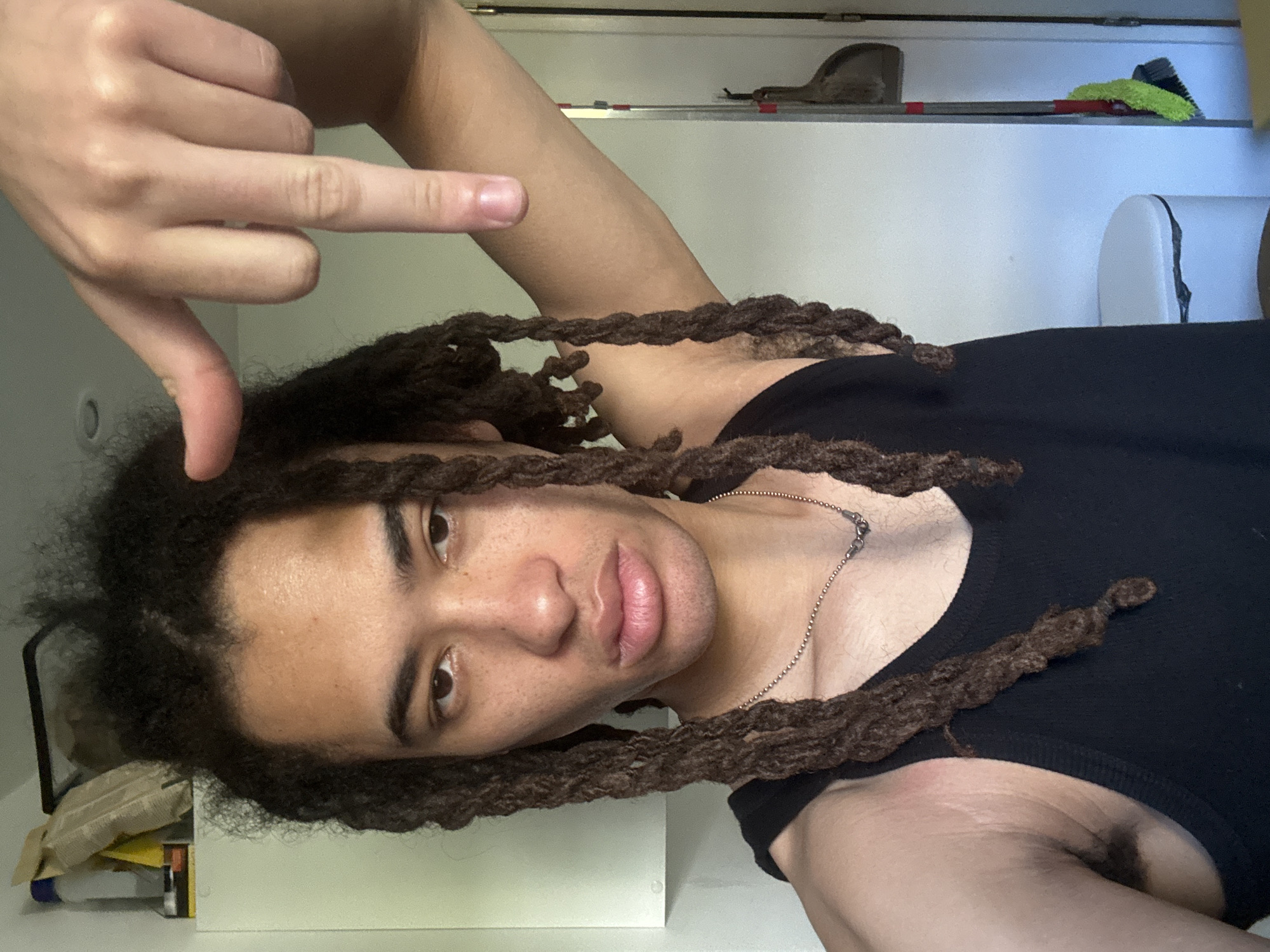 The image displays a person with light brown skin, dark eyes, and long, dark brown dreadlocks, wearing a black tank top and a silver ball-chain necklace. The photograph is oriented on its side, showing the person's head and upper torso. Their face is angled towards the viewer with a neutral expression, and their lips are slightly parted. Freckles are visible across their nose and cheeks.
The person's left hand is raised, forming a gesture where the thumb and pinky finger are extended, and the middle fingers are bent. This gesture is oriented downwards in the current image rotation. The upper part of their left arm and a portion of their armpit are visible.
The background shows a domestic indoor setting with white walls or shelving. On a shelf above the person's head (when upright), a grey dustpan and a green-bristled brush with a red handle are visible. Another cluttered shelf or surface is present to the person's left (below in the rotated image), holding items like a crumpled brown paper bag and yellow packaging. A white object, possibly a cushion or part of a chair, is partially seen in the upper right background.
The activity taking place is a close-up photograph or selfie. There is no discernible text within the image, and the time of day is indeterminate. No specific visual elements in the image confirm the location as Les Ulis, France.