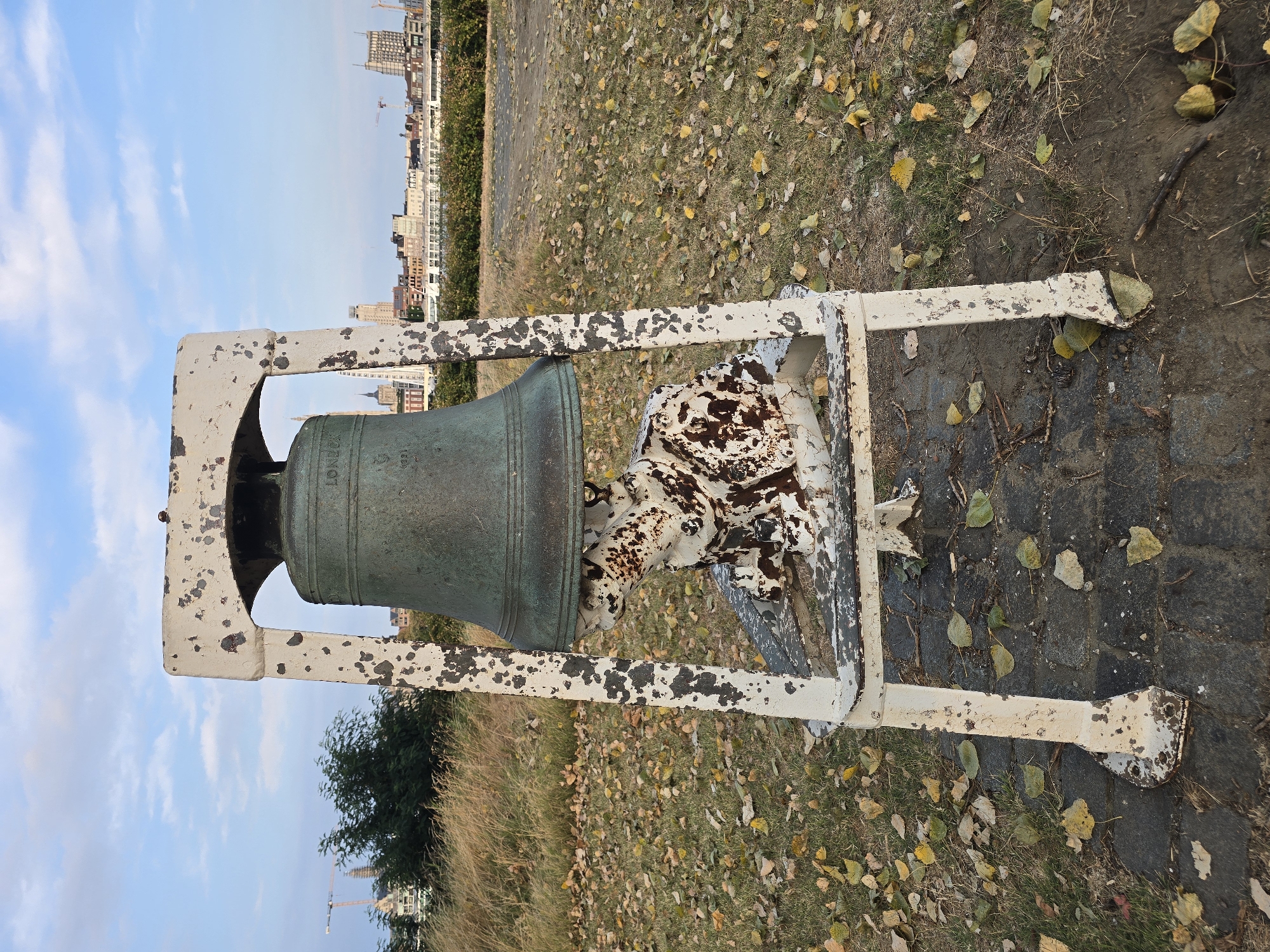 An outdoor, medium shot captures an old, verdigris-green bell mounted on a weathered, white metal A-frame structure. The bell has a maker's inscription, partially obscured, that appears to read "LONDON." The bell's stand is heavily distressed, with large patches of white paint chipped and peeling, revealing rusted metal underneath. At the base of the stand, a seated, possibly metallic, sculpture with a similar distressed, white and rust-colored finish is visible. The sculpture appears to be in a crouched or resting posture, with its form somewhat abstract.
The scene is set on a paved area transitioning into a grassy ground covered with fallen autumn leaves in shades of yellow and brown. In the background, a blurred cityscape with buildings and construction cranes is visible under a partly cloudy sky. The overall setting suggests an outdoor public space, possibly a park or a promenade along the water. The presence of the bell and sculpture indicates it might be a commemorative or artistic installation. The location is identified as Antwerp, Belgium, likely near the port or a waterfront area given the cityscape. The time of day appears to be late afternoon or early evening, with soft lighting. There are no people or animals present in the frame.