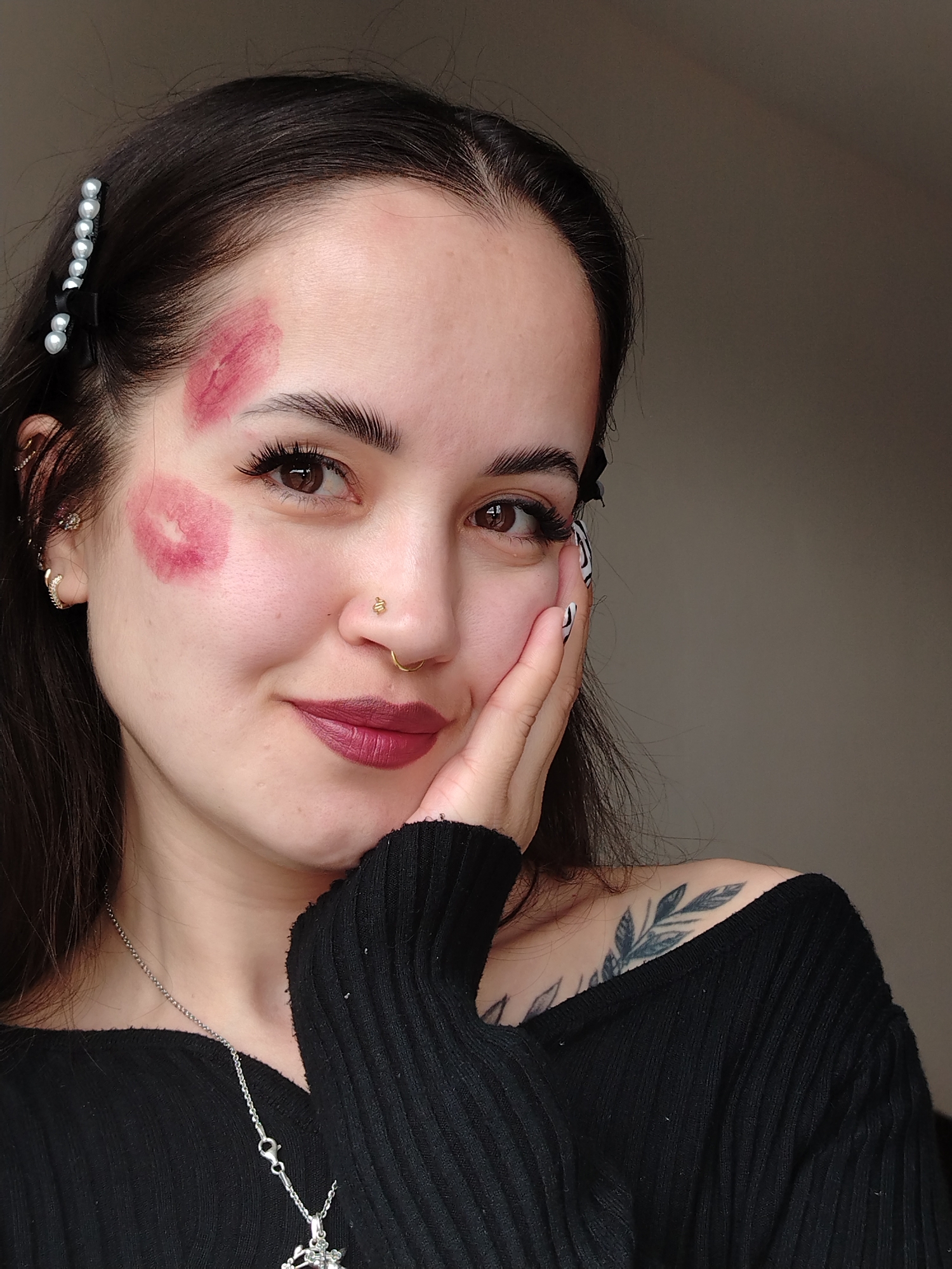 A young woman with dark hair is posing for a close-up selfie. She has fair skin with two lipstick kiss marks on her face: one on her forehead and one on her cheek. She is wearing a black off-the-shoulder sweater, a silver necklace, and has a floral tattoo on her left shoulder. A pearl and black ribbon hair clip adorns her hair. Her nails are painted with a black and white zebra-like pattern. The background is a plain light-colored wall. There is no indication that this photo was taken in Noviy, Russia.