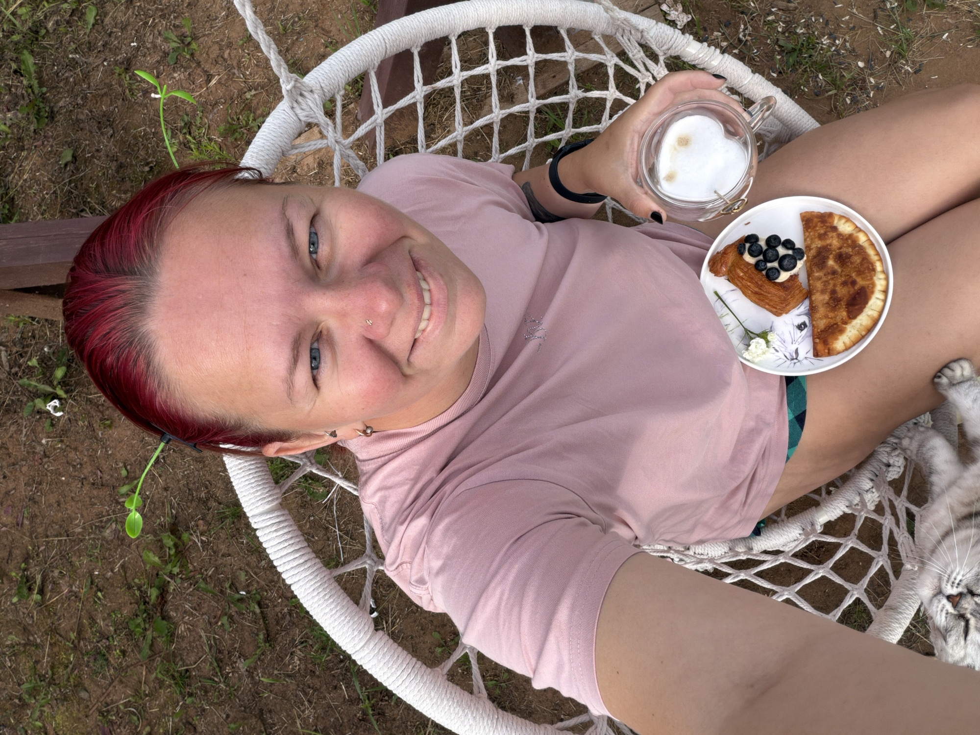A woman with bright red hair is sitting in a white macrame hanging chair outdoors, holding a plate with a pastry, blueberries, and a small fried pie, and a glass of coffee with latte art. She is wearing a pink t-shirt and green and black checkered shorts. A grey cat is lying on the ground at her feet. The background is a grassy, dirt area with some foliage.