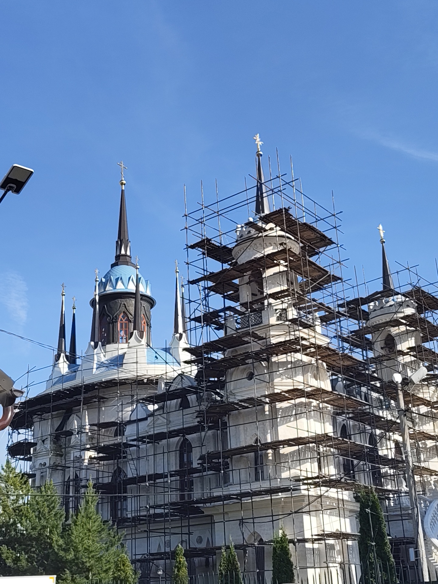 A low-angle, eye-level photograph captures the upper and mid-sections of the Church of the Vladimir Icon of the Mother of God in Bykovo, Moscow Oblast, Russia, which is currently undergoing renovation or construction. The large, ornate building features a predominantly white facade, characterized by multiple spires and domes. The most prominent central spire culminates in a distinctive multi-tiered dome structure composed of blue, white, and black sections, topped with a tall black conical spire and a golden Orthodox cross. Several other slender black spires, each topped with a golden Orthodox cross, are visible across the roofline. Extensive dark metal scaffolding, complete with wooden platforms, encases significant portions of the building, particularly the right half, obscuring parts of the white walls and windows. The sky overhead is clear and bright blue, occupying the upper two-thirds of the frame, with a faint, wispy cloud on the far left. Dark green coniferous trees are visible in the lower foreground, partially obscuring the base of the church. A modern, rectangular streetlight fixture on a dark pole extends into the frame from the far left. No individuals are discernible within the scene.