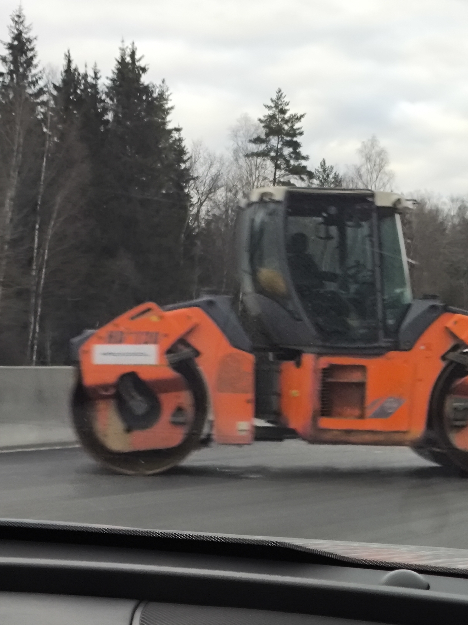 A construction roller, predominantly orange in color, is visible in the foreground. The machine has a black cabin and its wheels are blurred, suggesting it is in motion. Behind the roller, a grey concrete barrier is visible, separating the road from a wooded area. The wooded area consists of coniferous and deciduous trees, with the trees towards the back appearing darker due to the overcast sky. The photograph appears to have been taken from inside a vehicle. The location appears to be in Russia, but not the city of Kapan'. The city of Kapan' is located in Armenia.