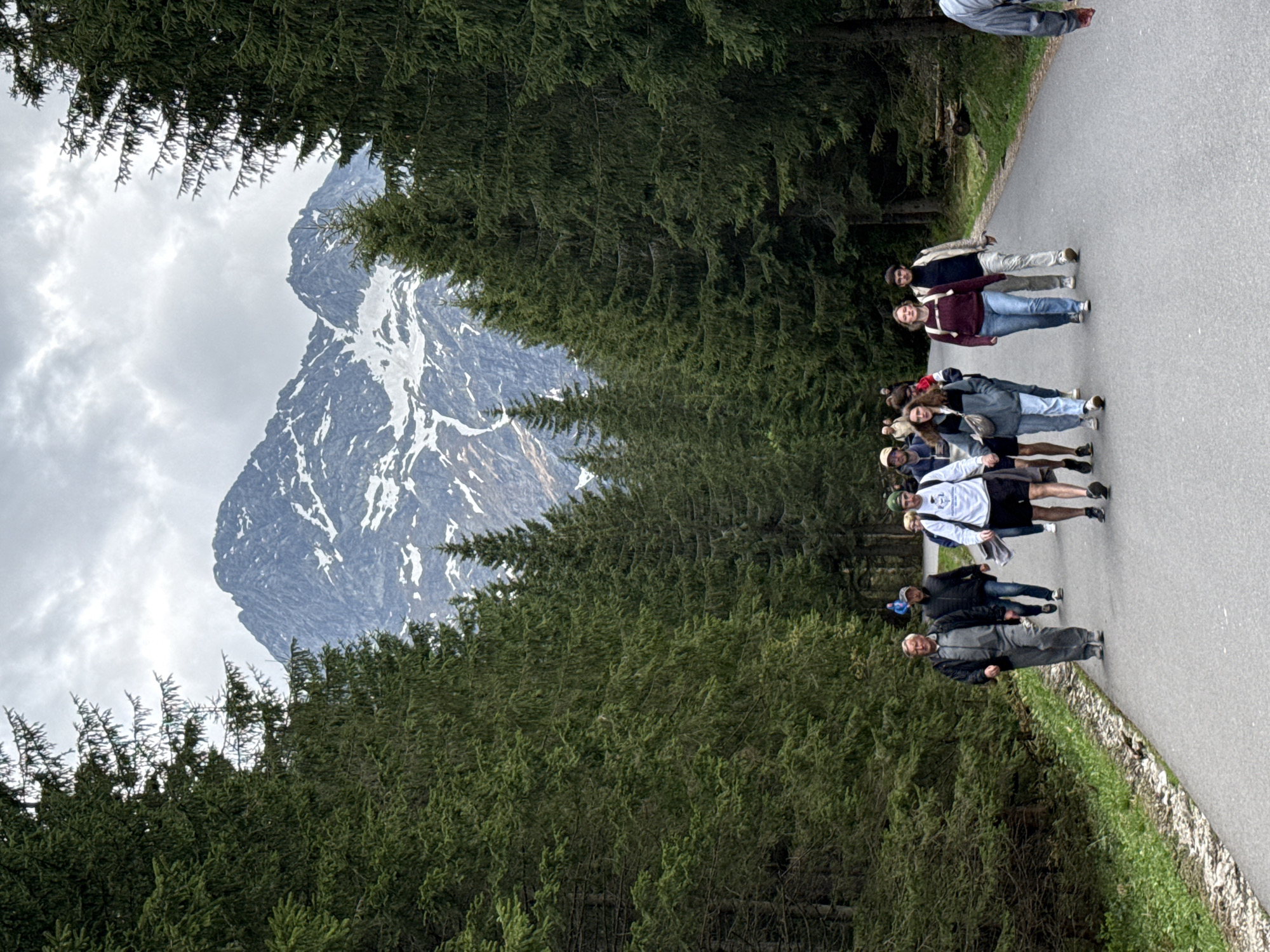 A group of approximately twelve people are walking along a paved road, next to a forest of evergreen trees. In the background, there is a tall mountain with visible snow patches. The sky is cloudy. The scene takes place in Brzegi, Poland.