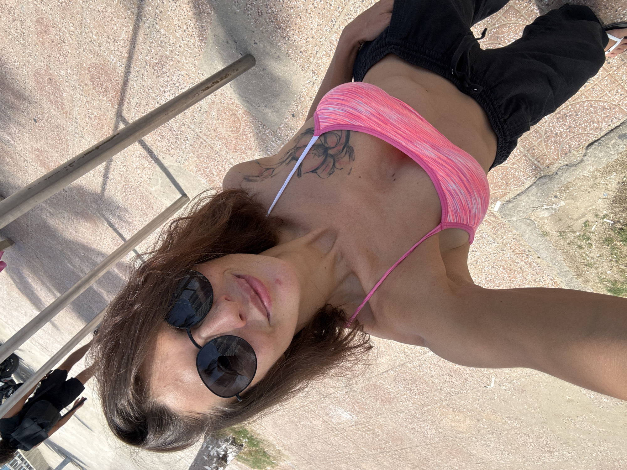 In Bac Nha Trang Ward, Vietnam, a woman stands outdoors on a paved walkway under sunlight. She is wearing a pink striped bikini top and black pants. The woman has brown hair, sunglasses, and a tattoo on her chest. To the left of the frame, another person wearing a black shirt and shorts with a backpack walks away from the camera, with a motorcycle visible in the background. The walkway appears to be made of textured concrete and decorative tiles.