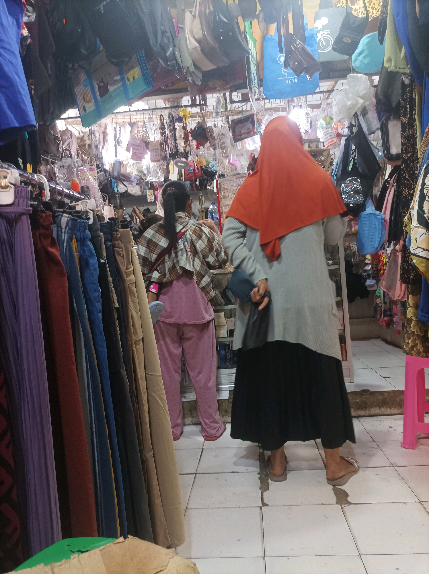 The image depicts an indoor market stall, likely in Sumuragung, Indonesia. In the foreground, several rows of vertically hung skirts and pants are displayed, ranging in color from purple to brown. To the right, a person wearing a gray long-sleeved sweater, a black skirt, and an orange hijab is standing. They are holding two dark-colored bags. To the left of this person, another individual is visible, wearing a plaid shirt and pink pants. This individual is facing away from the camera and appears to be browsing the merchandise. The stall is cluttered with various items, including bags, accessories, and other clothing, hanging from the ceiling and shelves. The flooring consists of white square tiles.