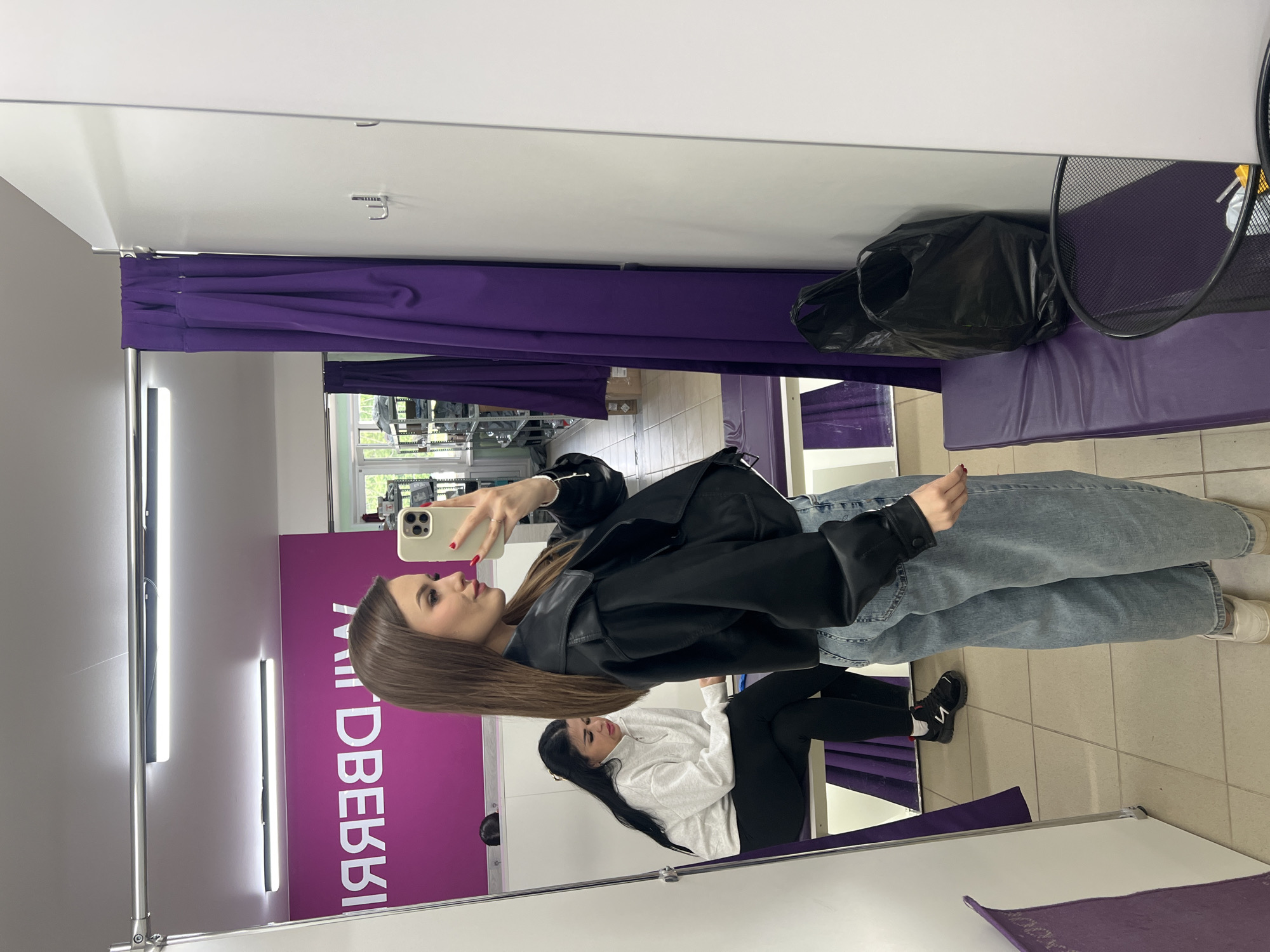 A young woman is seen in an indoor mirror selfie, standing and holding a light-colored smartphone to capture her image. She is wearing a black leather jacket over a top, light blue baggy jeans, and white sneakers. Her long, straight brown hair falls over her shoulders. Her reflection in the mirror shows her from a slightly different angle, revealing another person's reflection (or another part of her own reflection) seated behind her, wearing a white hoodie, black pants, and black sneakers.
The setting appears to be a fitting room or changing area within a retail establishment, characterized by purple curtains in the background and a large mirror. Fluorescent light fixtures are visible on the ceiling, and the floor is made of light-colored tiles. Hooks are attached to the ceiling above the curtains. To the right of the main subject, a black plastic bag and a black wire mesh basket are visible, likely placed on a purple bench or shelf.
A notable detail is the mirrored text "ЯЛБВЕЯЯЛ" visible on the purple wall behind the reflection, which spells "Вайлдберриз" (Wildberries) backward. This indicates the photo was taken at a Wildberries store or pickup point, located in Noviy, Russia. The woman's expression is neutral as she takes the selfie. The time of day and weather are not discernible from the indoor scene with artificial lighting.