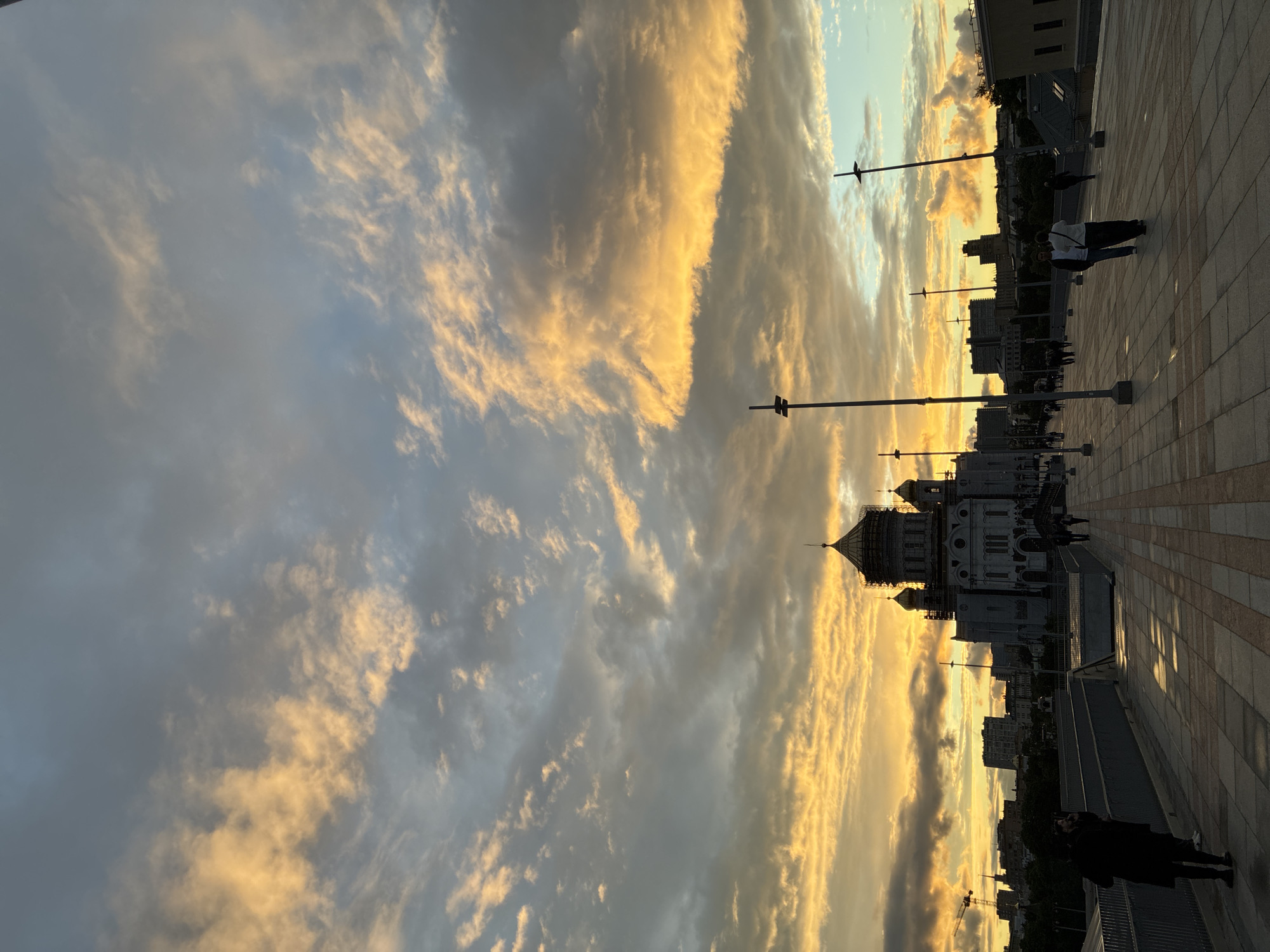 The image shows the Cathedral of Christ the Savior in Moscow, Russia, at sunset. The sky is filled with clouds, some of which are illuminated in golden light. The cathedral is white with a golden dome. People are walking along the pavement in front of the cathedral. Tall poles with lights are lined up along the walkway. Buildings are visible in the background.