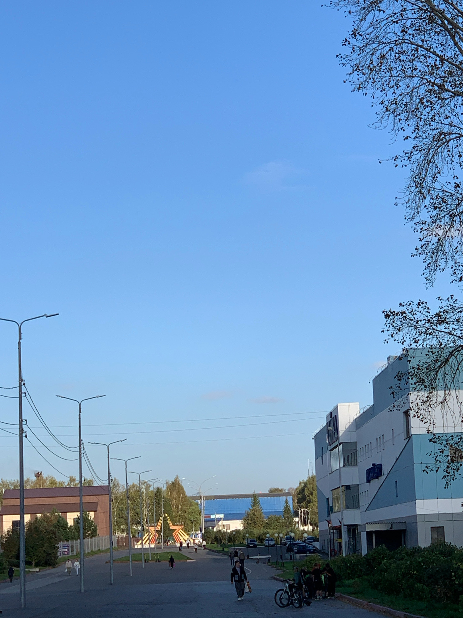 The image depicts a street scene in Prokopyevsk, Russia, on a clear day with a bright blue sky. In the foreground, a paved area with several streetlights is visible. A group of people are walking on the paved area, and a few are standing or gathered in groups. One person is walking a bicycle. To the right, a modern building with a light blue and white facade is prominent. In the middle ground, a playground structure with two large red and yellow star shapes is present, along with trees and other buildings. In the background, more trees and buildings are visible. The overall impression is of an urban environment with public spaces and commercial structures.