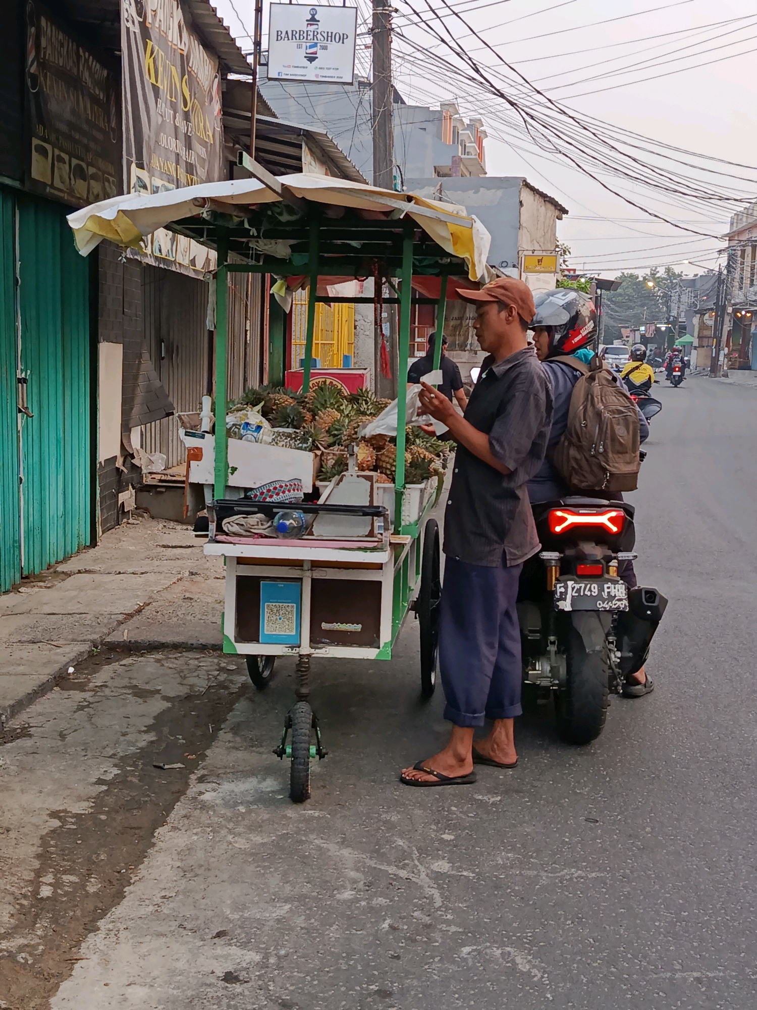An outdoor scene in Bekasi, Indonesia, captures a street vendor selling pineapples from a cart on the roadside. The vendor, a man wearing a brown cap, dark grey shirt, and blue pants, stands beside the cart, holding a white plastic bag. Two individuals are on a motorcycle parked behind the cart; the passenger, wearing a helmet, has a brown backpack. The cart, with a yellow and white awning, is laden with pineapples. To the left of the cart, a barbershop with a sign displaying "BARBERSHOP EST. 2000" is visible. The street is paved, with a wet patch on the left side. Electrical wires crisscross overhead. In the background, buildings and more street activity, including other motorcycles, are present.