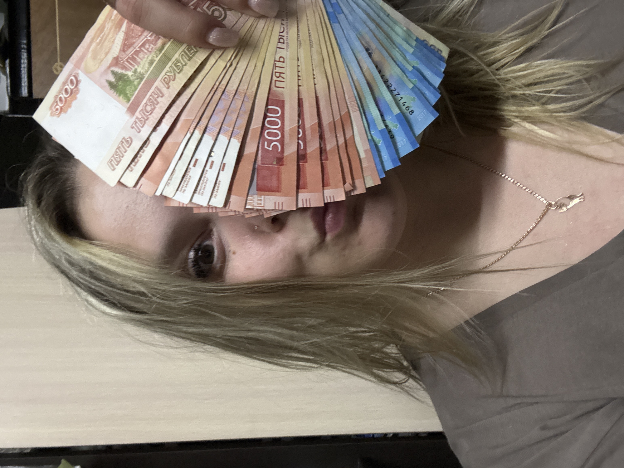 A woman poses with a fan of Russian 5000 ruble banknotes held in front of her face. The notes are mostly red and yellow, with some blue ones visible at the edge. The woman has blonde hair and is wearing a grey shirt and a gold necklace. Only her eye, nose, and mouth are visible behind the money. The location is unknown and there is no indication of the city of Markovskiy, Russia in the image.