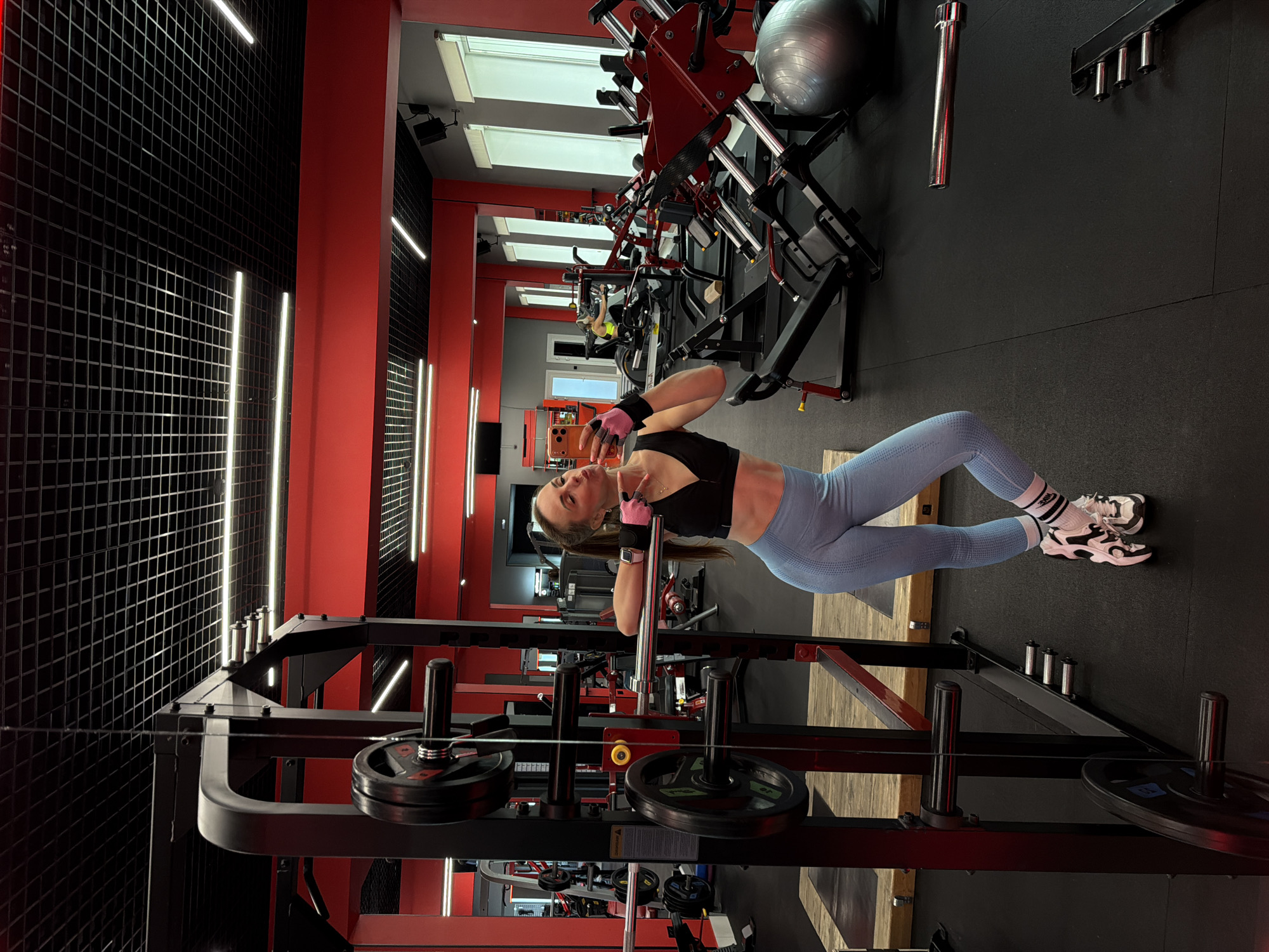 A woman with long brown hair pulled into a ponytail is taking a selfie in a gym. She is wearing a black sports bra, light blue textured leggings, white athletic socks with black stripes, and black and white athletic shoes. She has pink workout gloves on her hands. She is leaning against a weight rack with weight plates. In the background, there are various gym machines, including leg press machines and a rowing machine. The gym has red walls and a black ceiling with grid lighting. There are windows with white blinds on the right side of the image. The scene is indoors in a modern gym. The location is specified as Birobidzhan, Russia.