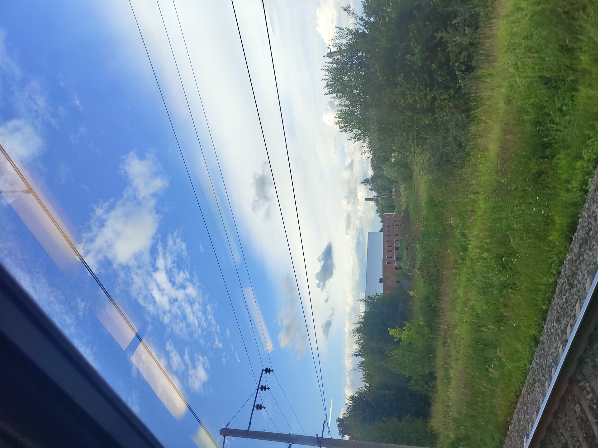 The image shows a view from a train window. In the foreground, there are train tracks. The middle ground is a grassy field with trees. In the background, there is a building under construction with a white roof and brick walls. There are also power lines stretching across the sky. The sky is blue with white clouds. The image appears to have been taken in Фруктовая, Russia.