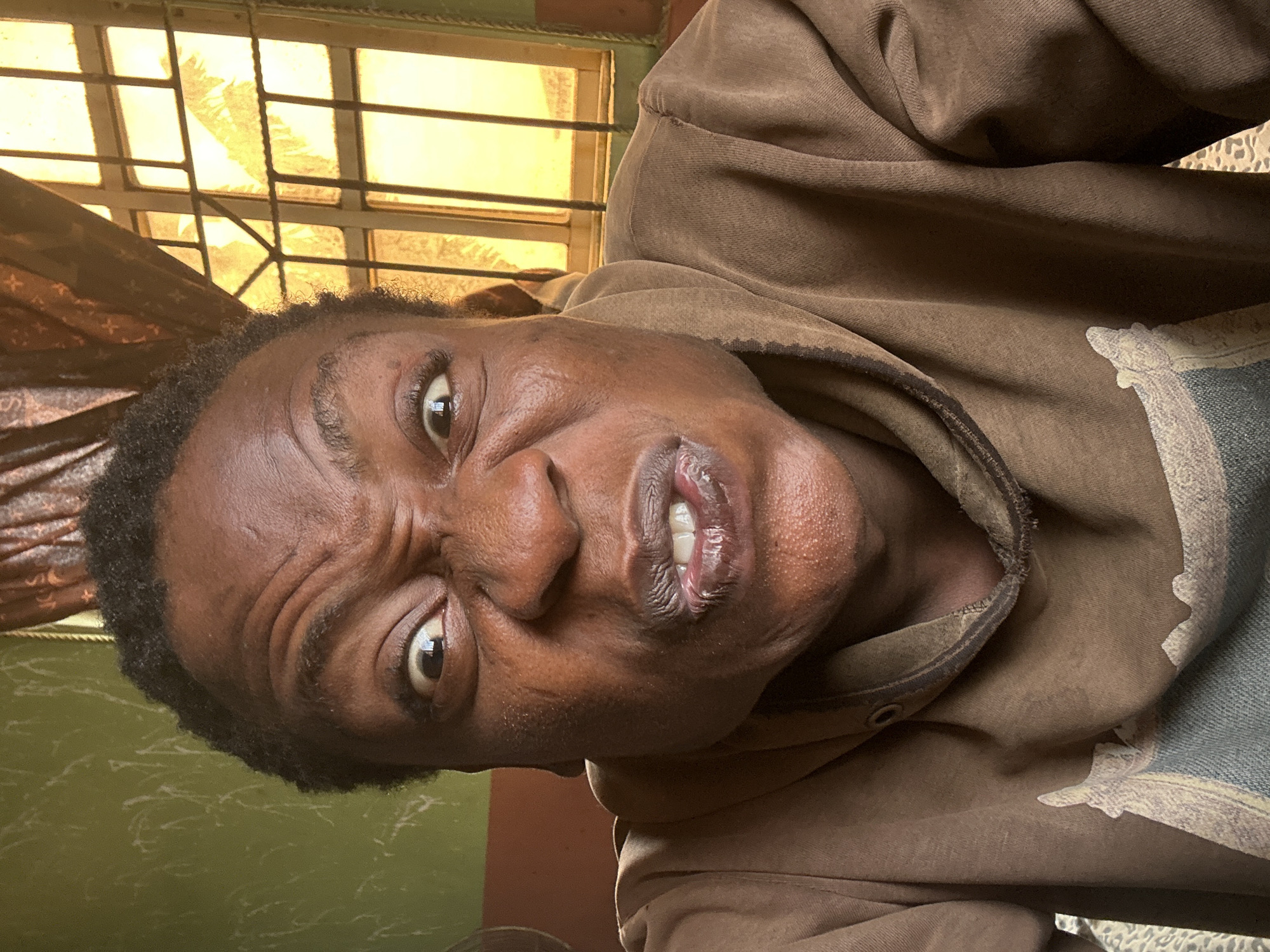 A close-up, tilted shot of a person's face wearing a brown hooded sweatshirt. The person has dark skin, short black hair, and a contorted facial expression with furrowed brows. The background shows a window with a grid-like pattern. The location is Unknown, Unknown.