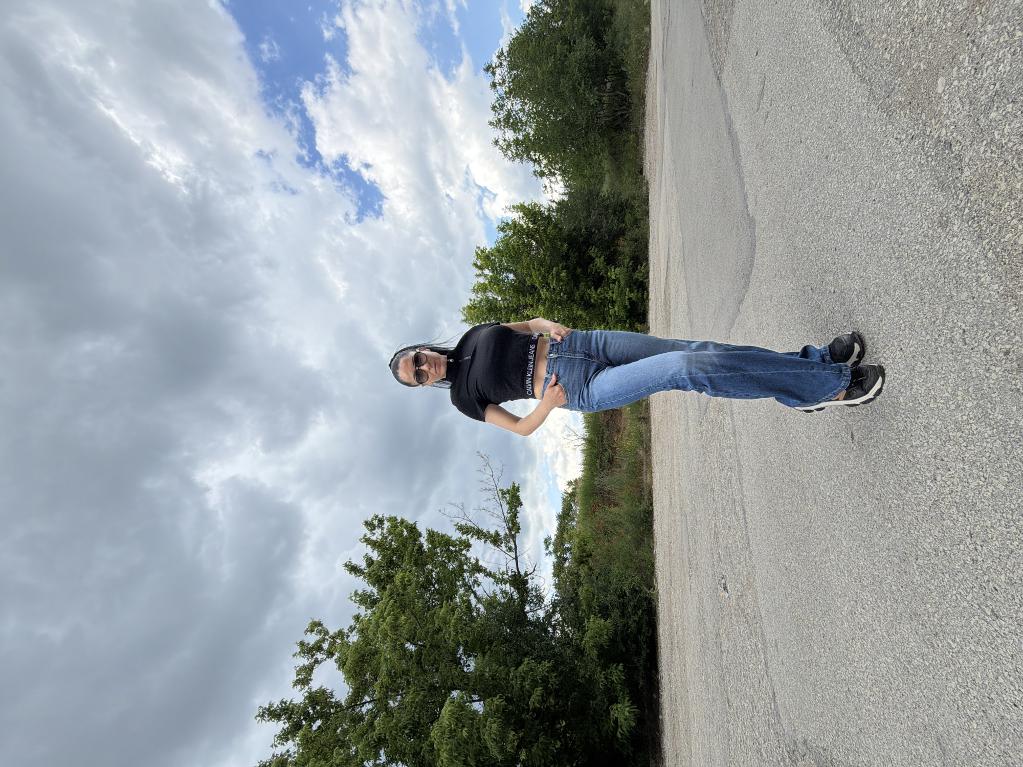 A full shot of a woman in sunglasses standing on an asphalt road. She is wearing a black crop top with white letters on the band, blue jeans, and dark sneakers with white soles. She has dark hair and is holding her hands on her hips. The road is gray and stretches into the distance. On either side of the road is green vegetation, including trees and bushes. The sky above is cloudy with patches of blue. The image is vertical. The location appears to be somewhere outside the city of Pleven, Bulgaria due to the surrounding nature and road.