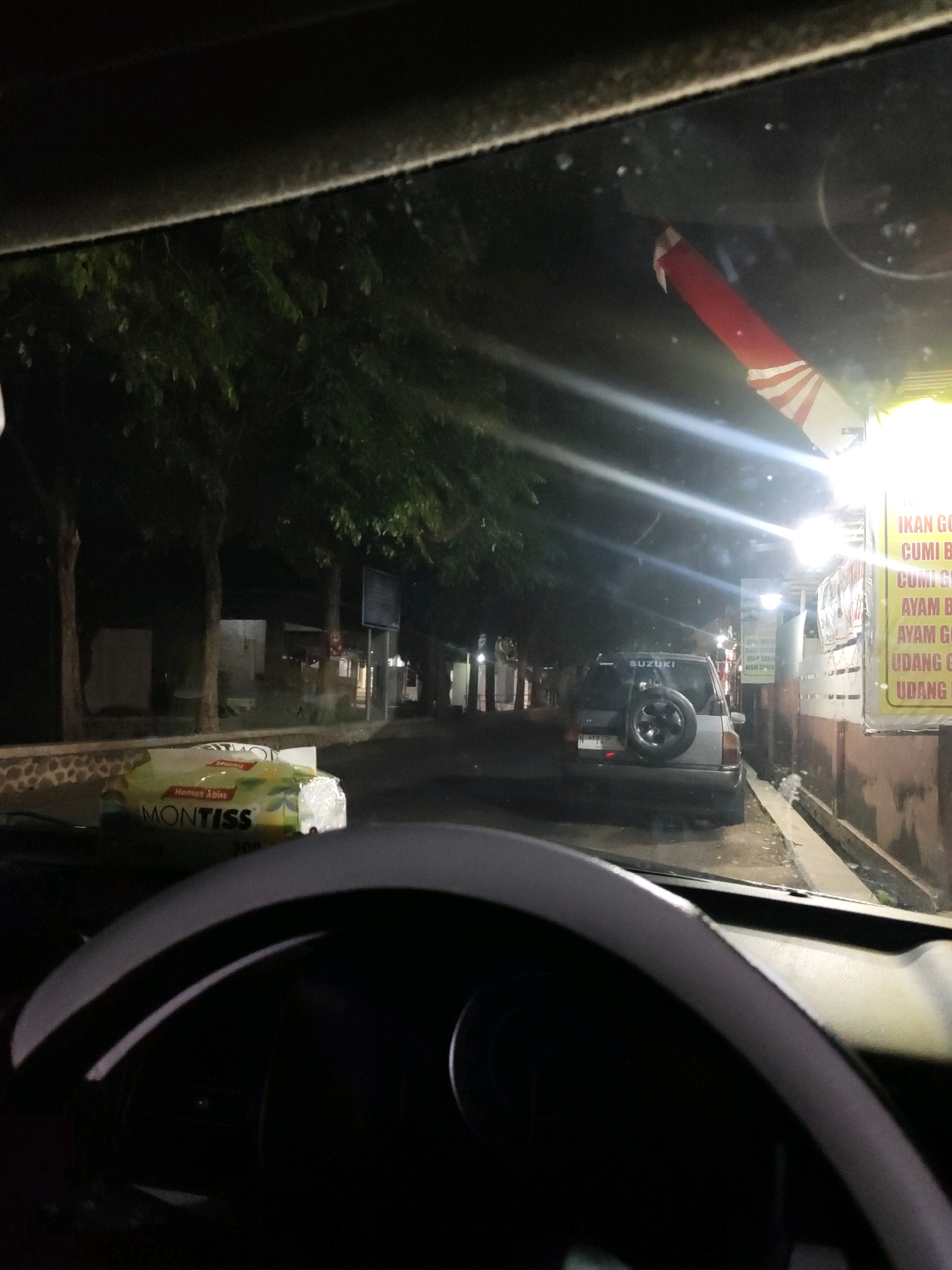 The image depicts a nighttime scene from inside a vehicle, looking out through the windshield. In the foreground, a portion of the car's steering wheel is visible. On the dashboard, a pack of Montiss tissues is prominently displayed.
The exterior environment shows a street lined with trees. Several buildings are present, one of which has a bright sign with text that appears to be in Indonesian, listing food items such as "IKAN GORENG" (fried fish), "CUMI BUMBU" (squid dish), "AYAM BAKAR" (grilled chicken), and "UDANG GORENG" (fried shrimp). Another building shows a sign with the brand "SUZUKI" on a vehicle parked on the side of the road, which is a grey SUV with a spare tire mounted on the back. The road surface appears wet, suggesting recent rain.
A decorative item with red and white stripes, resembling a Japanese flag or a firework, is affixed to the windshield from the inside, near the top edge. The lighting is artificial, coming from streetlights and the illuminated signs of the establishments. The overall atmosphere is one of a commercial street in Probolinggo, Indonesia, at night.