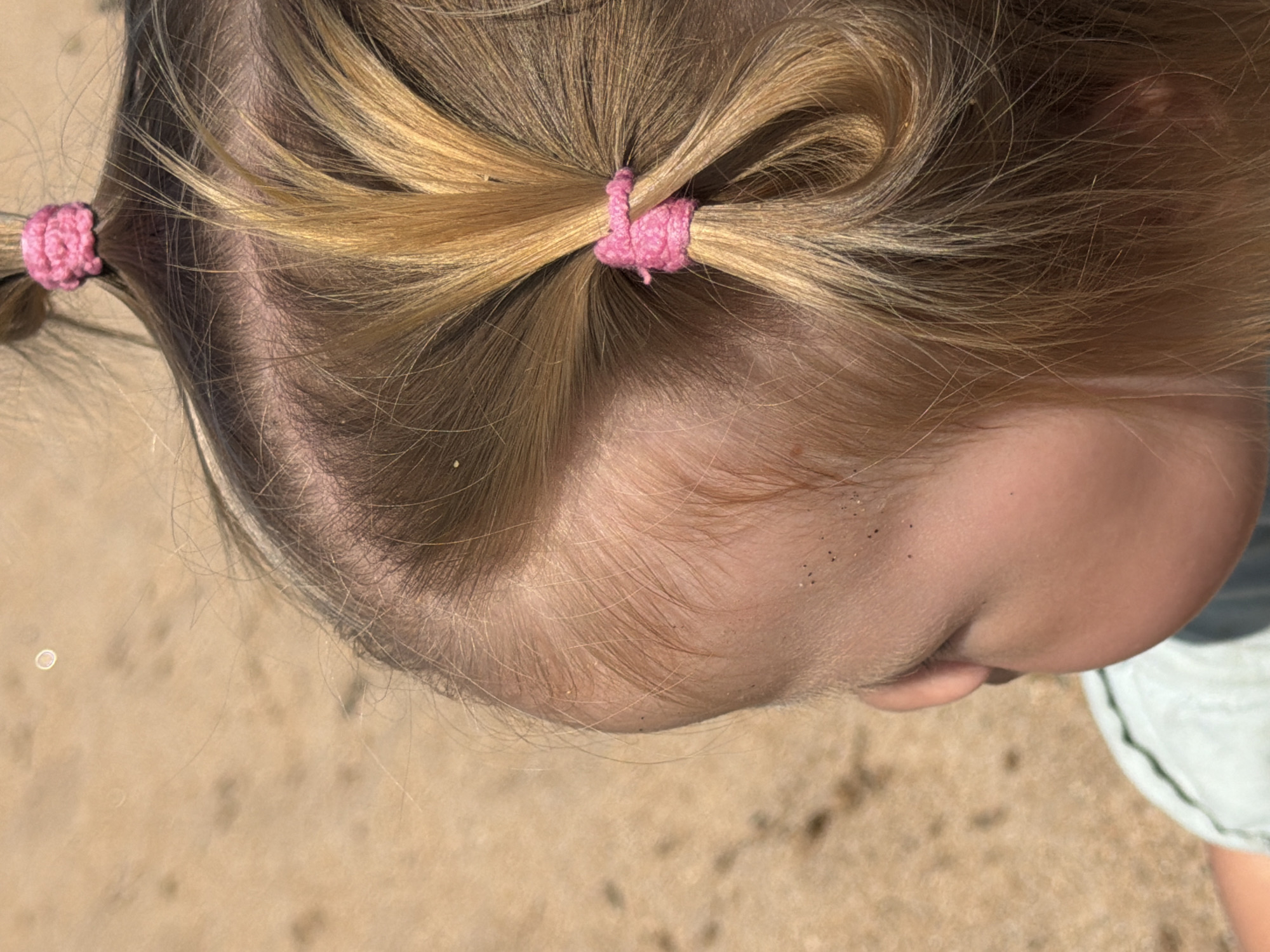 A close-up shot captures a young child's head from a high angle. The child's blonde hair is styled into two pigtails, each secured with a pink crocheted hair tie. The child's profile is visible, facing left, with a portion of their cheek and ear showing. The background consists of sandy ground, with small specks of dirt or sand visible on the child's skin and hair. The scene appears to be outdoors, possibly at a beach or a sandy area in Maksimiha, Russia.
