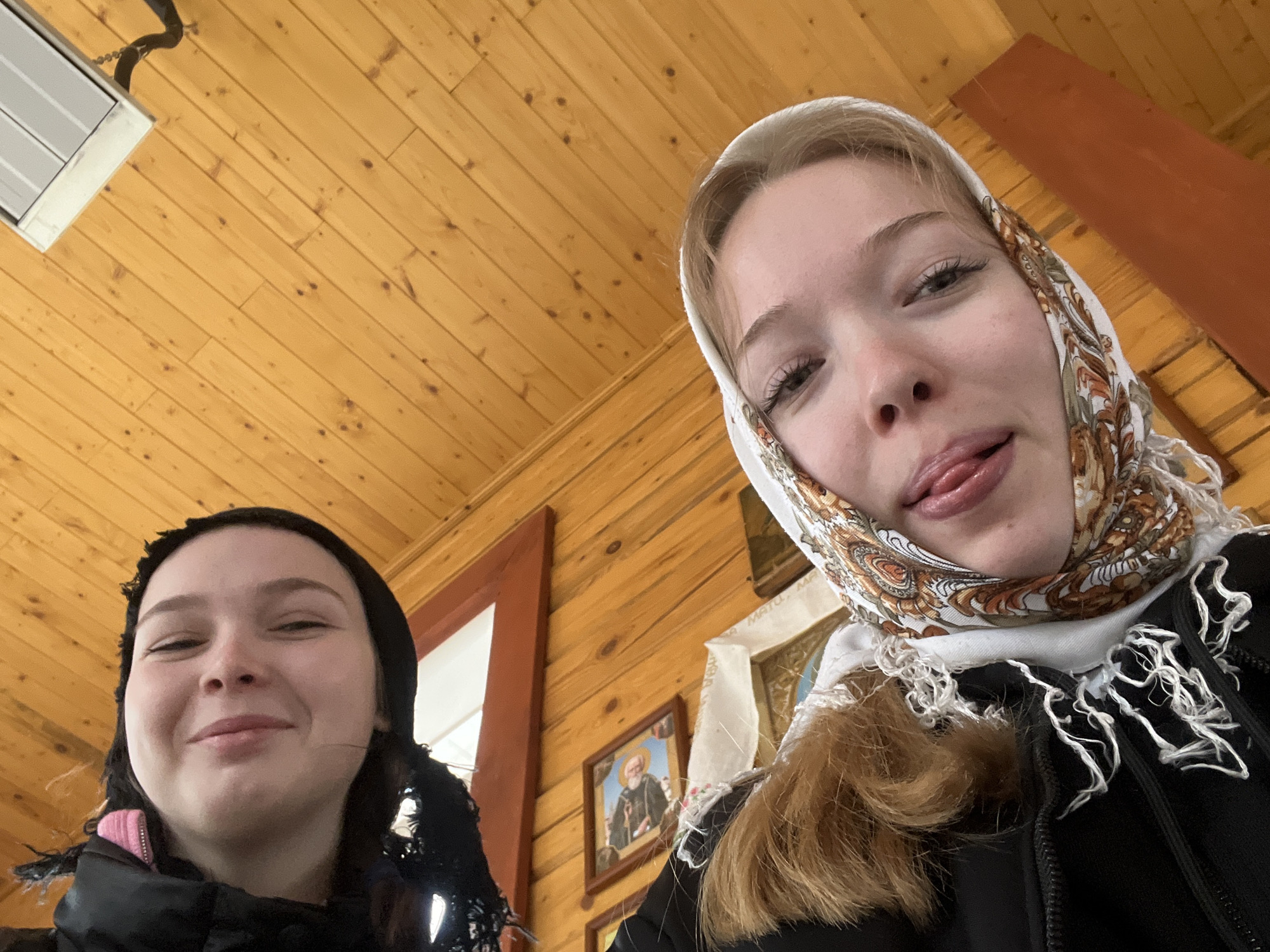 Two young women are in a wooden-walled room. The woman on the left has dark hair covered with a black head covering. The woman on the right has blonde hair and is wearing a white head covering with a brown and gold pattern. She is sticking her tongue out. In the background are wooden walls and framed religious icons. The location is indicated as Сосново, Russia.