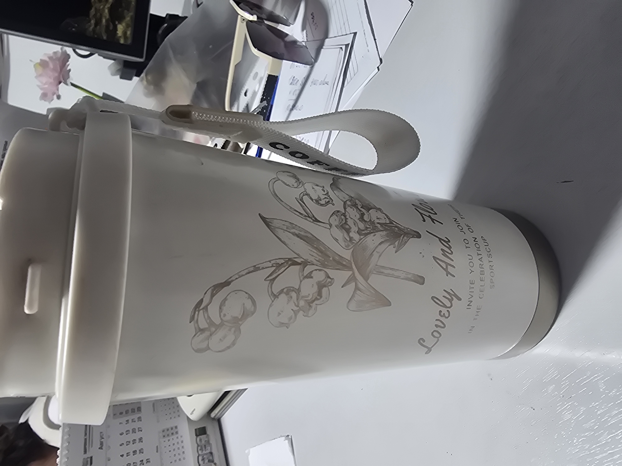 A white thermos with a lily of the valley design is placed on a white desk. The thermos has a beige lid and a white fabric loop attached to the side. The loop has black lettering that reads "COFFEE". Text on the thermos reads "Lovely And Flow" with smaller text below reading "INVITE YOU TO JOIN IN THE CELEBRATION OF THEIR SPORTSCUP". In the background, a calendar is visible, along with a pink flower in a vase, and a pair of sunglasses on some papers. The scene appears to be an indoor setting, possibly an office or study. There is no information in the image to suggest a location in Ulan-Ude, Russia.