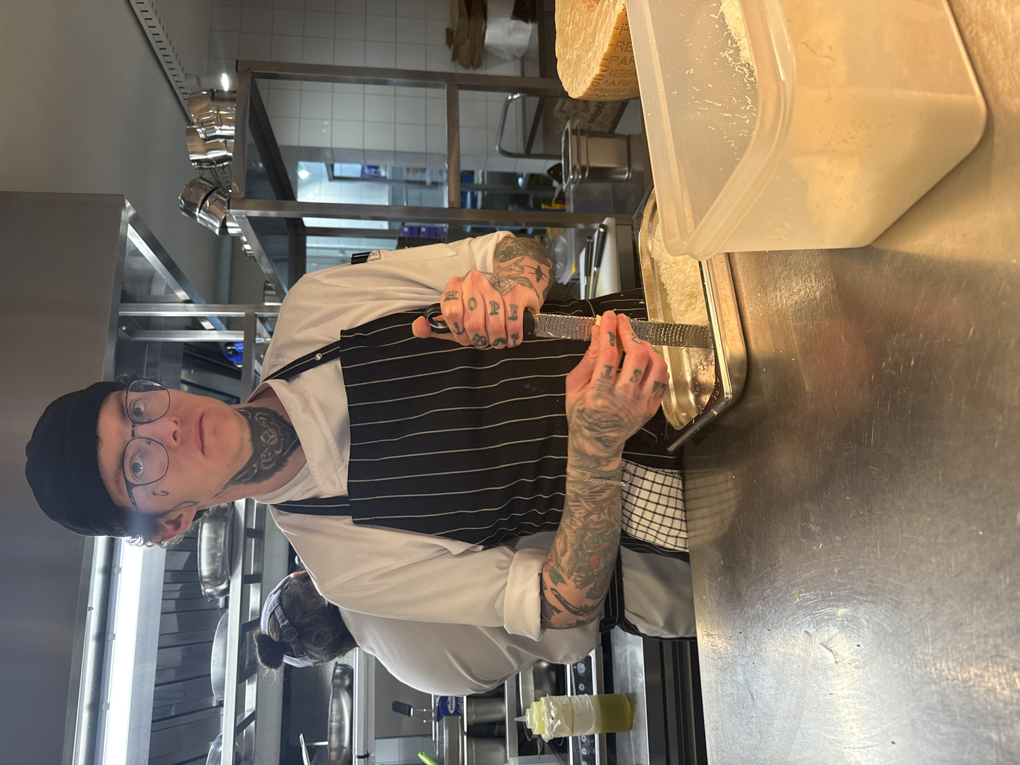 A chef with tattoos on his hands and neck, wearing a black beanie, round glasses, a white chef's jacket, and a black-and-white striped apron, is grating cheese into a clear plastic container. He is positioned at a stainless steel counter in a kitchen setting. Behind him, pots and pans hang from a rack. To his right, a wheel of cheese is visible. The location is Unknown, Unknown.