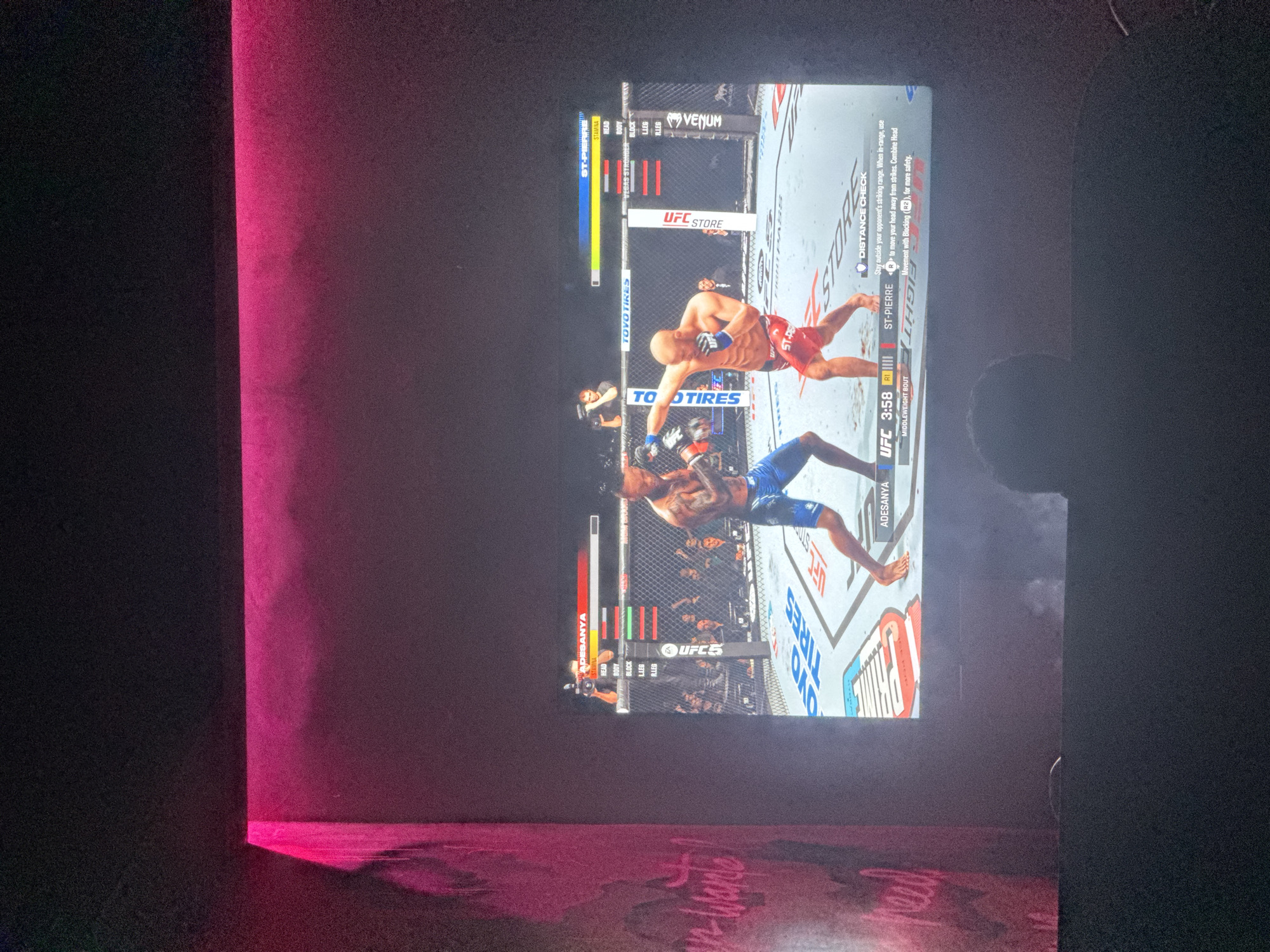 A screen displays the UFC 5 video game, with the match between Adesanya in blue shorts and St-Pierre in red shorts. The fight is in the first round, 3:58 left. The screen is in a dimly lit room, partially illuminated by a pink light. Some graffiti is visible below the screen. The location in the city of Kuta Selatan, Indonesia is not discernible from the visual details in the image.