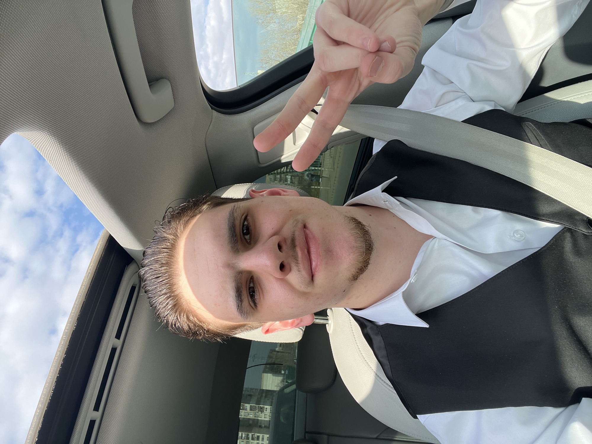 A young adult male is seated inside a vehicle, capturing a self-portrait. He has slicked-back dark hair, a goatee, and is wearing a white collared shirt beneath a black vest. A light grey seatbelt is visible across his right shoulder and chest. His right hand is raised, displaying a "peace" sign with the index and middle fingers extended. A prominent shadow from an overhead light source diagonally traverses his forehead and hair. The vehicle's interior features light-colored upholstery and a visible open sunroof, revealing a partly cloudy blue sky. Through the rear window on the left, a multi-story residential building with numerous windows, characteristic of urban apartment blocks in Sofia, Bulgaria, is visible. The passenger side window on the right reveals the sky, distant foliage, and a low green barrier, indicative of an urban environment under clear daylight conditions.