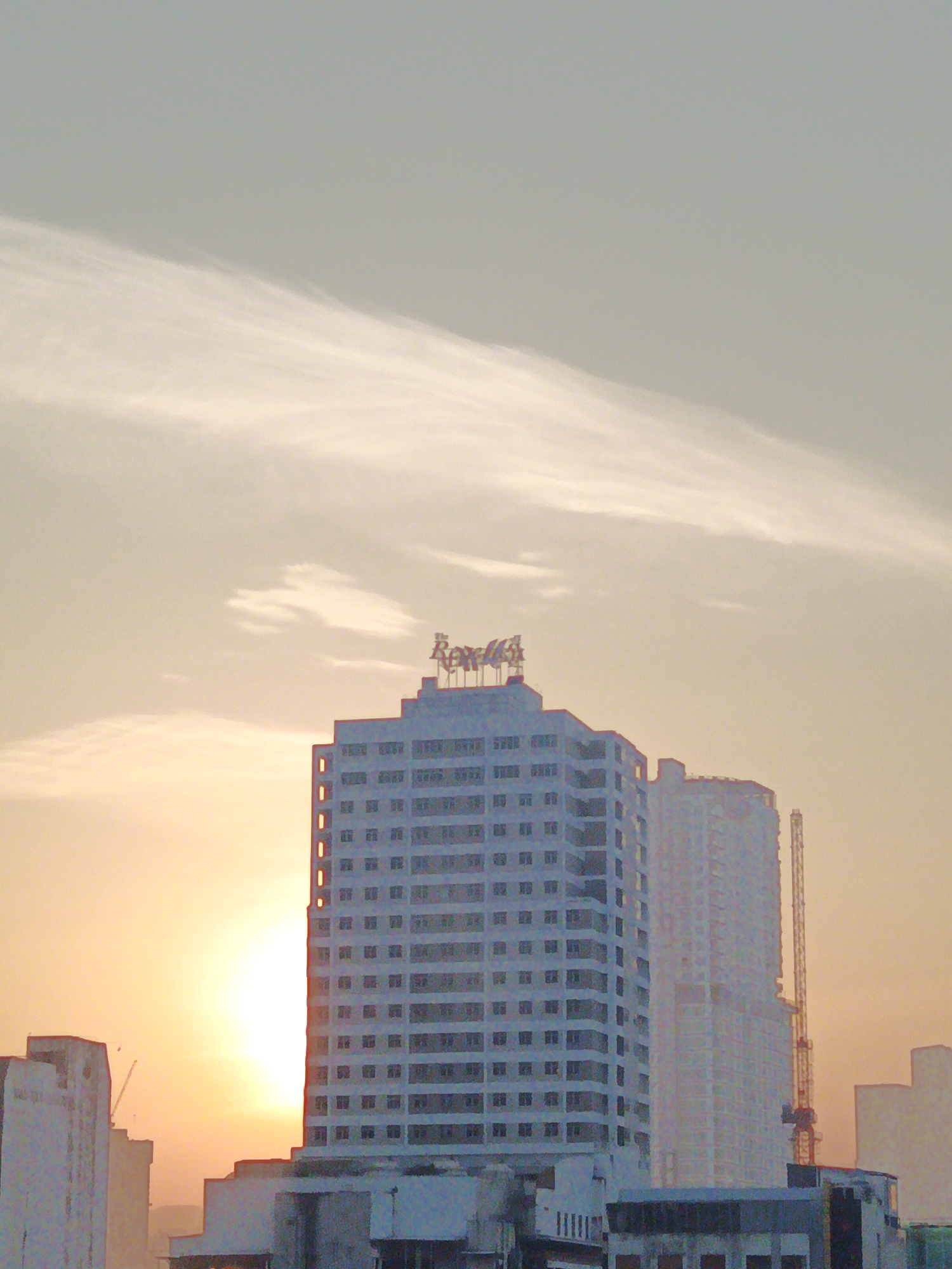 The image captures a city skyline at sunset, featuring several tall buildings and a crane. The sky is a gradient of orange and light blue, with wispy clouds. The most prominent building in the foreground is a white, multi-story structure with numerous windows and a sign at the top that appears to read "Royale." To the right of this building, another tall, lighter-colored building is under construction, with a construction crane extending upwards next to it. In the background, more buildings are visible, with the setting sun casting a warm glow on the scene. The overall impression is that of an urban environment during twilight.