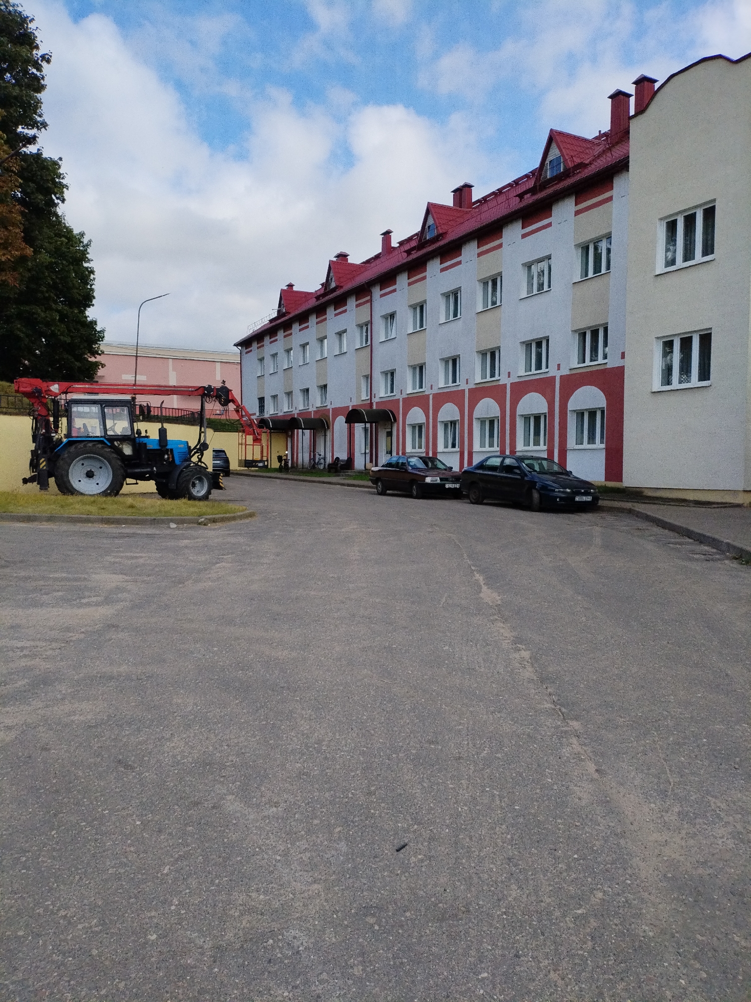 The image depicts a street scene in Bialynichy, Belarus, featuring a multi-story building with a red roof and a pale facade accented with red horizontal stripes and white window frames. In front of the building, two dark-colored sedans are parked on the asphalt. To the left, a blue tractor with a red crane attachment is situated on a patch of grass. A tall street lamp stands to the left of the tractor. The sky above is partially cloudy with a blue hue. The foreground consists of an asphalt road.
