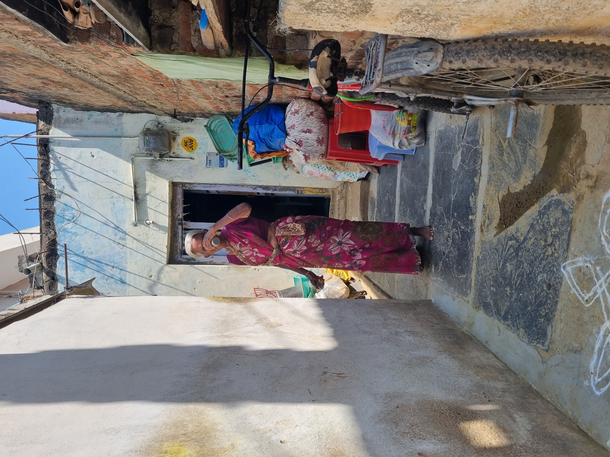 An elderly woman stands in a narrow, sunlit alleyway in Pedda Kammavaripalli, India. She is barefoot, dressed in a dark pink floral-patterned sari, and holding a metallic cup to her mouth, appearing to drink. Her white hair is pulled back, and her gaze is directed slightly to her left. The alley is bordered by a light-colored, textured wall on the left, casting a sharp shadow. On the right, a distressed light blue wall with an open dark doorway is visible, featuring an electrical meter box labeled "HSC 5008" and "159." Above, exposed rebar and electrical wiring are present. Adjacent to the doorway, various household items are stacked, including a green basket, a blue bag, a white floral bag, a red plastic stool, a red bucket, and a light blue stool with a plastic bag of colorful items. A dark-colored bicycle with a worn black and white seat leans against a reddish-brown brick wall in the immediate foreground on the right, its rear wheel showing dirt. The ground consists of concrete and dark flagstones, with white chalk floor art (Kolam) in the foreground and a visible damp patch. A clear blue sky is visible above the structures.