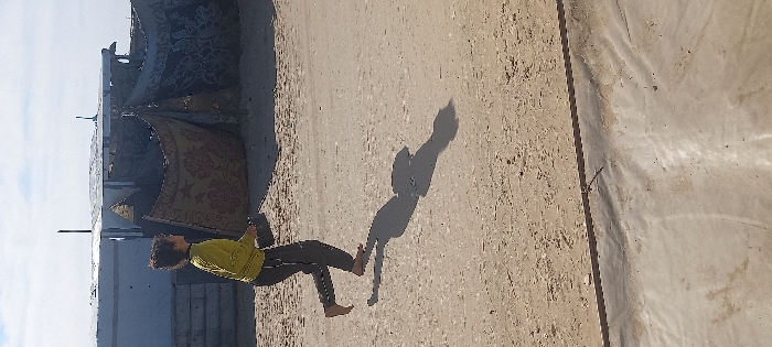 This image, taken in Khan Yunis, Palestinian Territory, depicts a daytime outdoor scene. A young, barefoot individual with dark, curly hair, wearing a long-sleeved yellow shirt and dark trousers, is shown mid-stride on a light brown, sandy ground, moving towards the right side of the frame. The individual holds a dark, rectangular object in their left hand. A distinct shadow of the individual is cast behind and to their right on the ground. In the background, a series of low-lying, improvised structures are visible, constructed from various materials including corrugated metal panels, tarpaulins, and patterned fabric, resembling rugs or blankets, used as walls or coverings. Dark, slender vertical elements, possibly poles, extend above these structures. The upper portion of the image shows a light blue sky with sparse white clouds. A dark, thin linear element is present horizontally across the bottom section of the foreground.